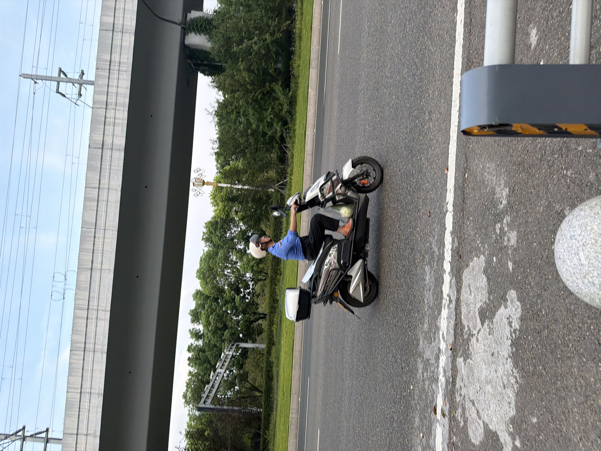 In Jiangjin, China, a person wearing a blue shirt, black pants, and a white helmet rides a silver electric scooter on an asphalt road. The scooter has a black storage box on the rear. Behind the scooter, a concrete overpass supported by pillars is visible, with power lines running above it. Lush green trees and vegetation line the side of the road, and a white line marks the edge of the lane. The sky is overcast with a pale blue hue. In the foreground, a rounded grey object and a dark grey barrier with yellow markings are partially visible.