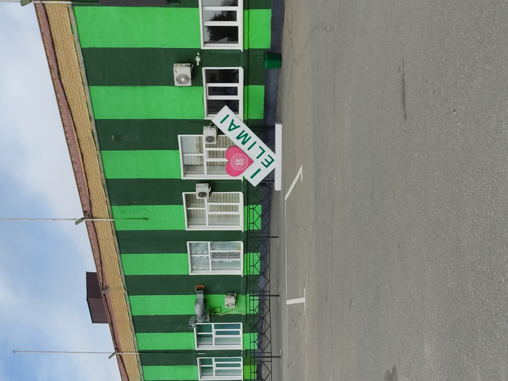 In Semey, Kazakhstan, an exterior shot captures the facade of a building painted with alternating dark green and bright green vertical stripes. The building has a beige brick-like material along the top and a darker brown, sloped roof section visible. Several white-framed windows are present, some appearing to have blinds. An air conditioning unit is mounted on the exterior wall near the right side of the frame, and another is visible to the left of the "I ELIMAI" sign. A black metal fence runs along the base of the building, separating it from a paved area. In the foreground, a large, white, three-dimensional sign with a pink heart and the words "I ELIMAI" diagonally printed on it is positioned. The heart has a circular emblem with what appears to be a stylized 'G' inside. A green trash receptacle stands to the right of the sign. The ground is a grey asphalt surface, marked with white lines for parking spaces. The sky above is partly cloudy.