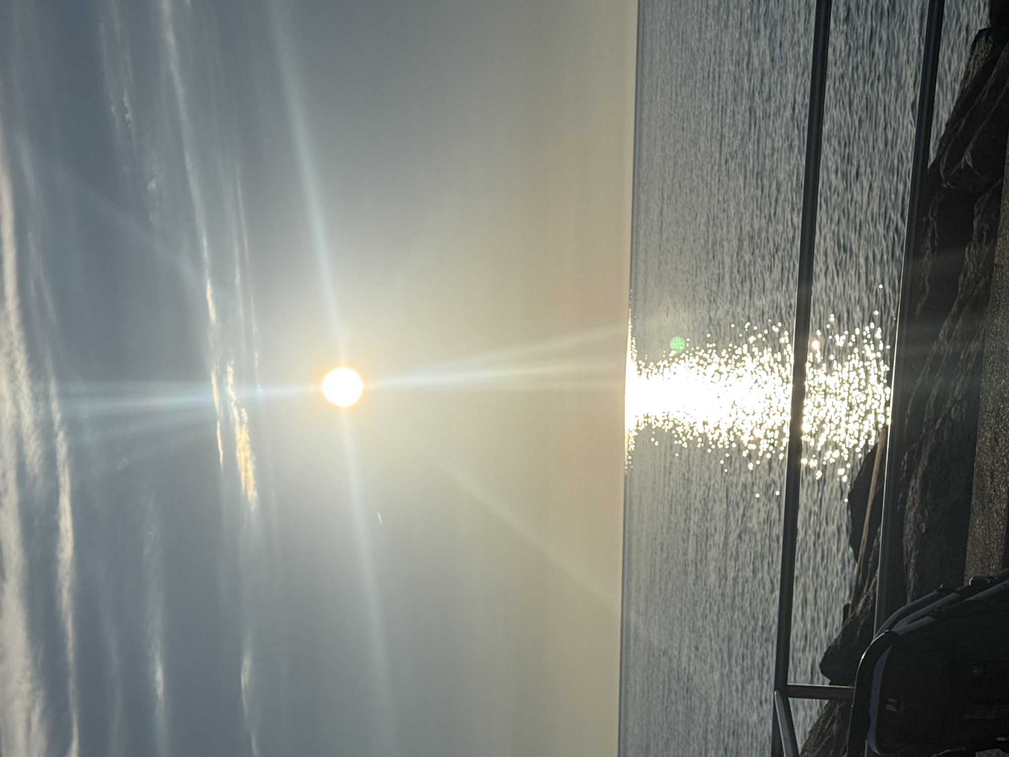 A bright sun shines in the upper center of the image, casting a brilliant reflection on the calm sea below. The sun is surrounded by lens flare, with rays extending outwards. Wispy clouds are scattered across the sky. A dark railing runs horizontally across the lower part of the image, separating the viewer from the rocky shore and the sea. In the lower left corner, a dark backpack or bag rests on the rocky ground. The scene appears to be a scenic viewpoint overlooking the Black Sea in Batumi, Georgia, likely during sunrise or sunset given the low angle of the sun and the soft light.