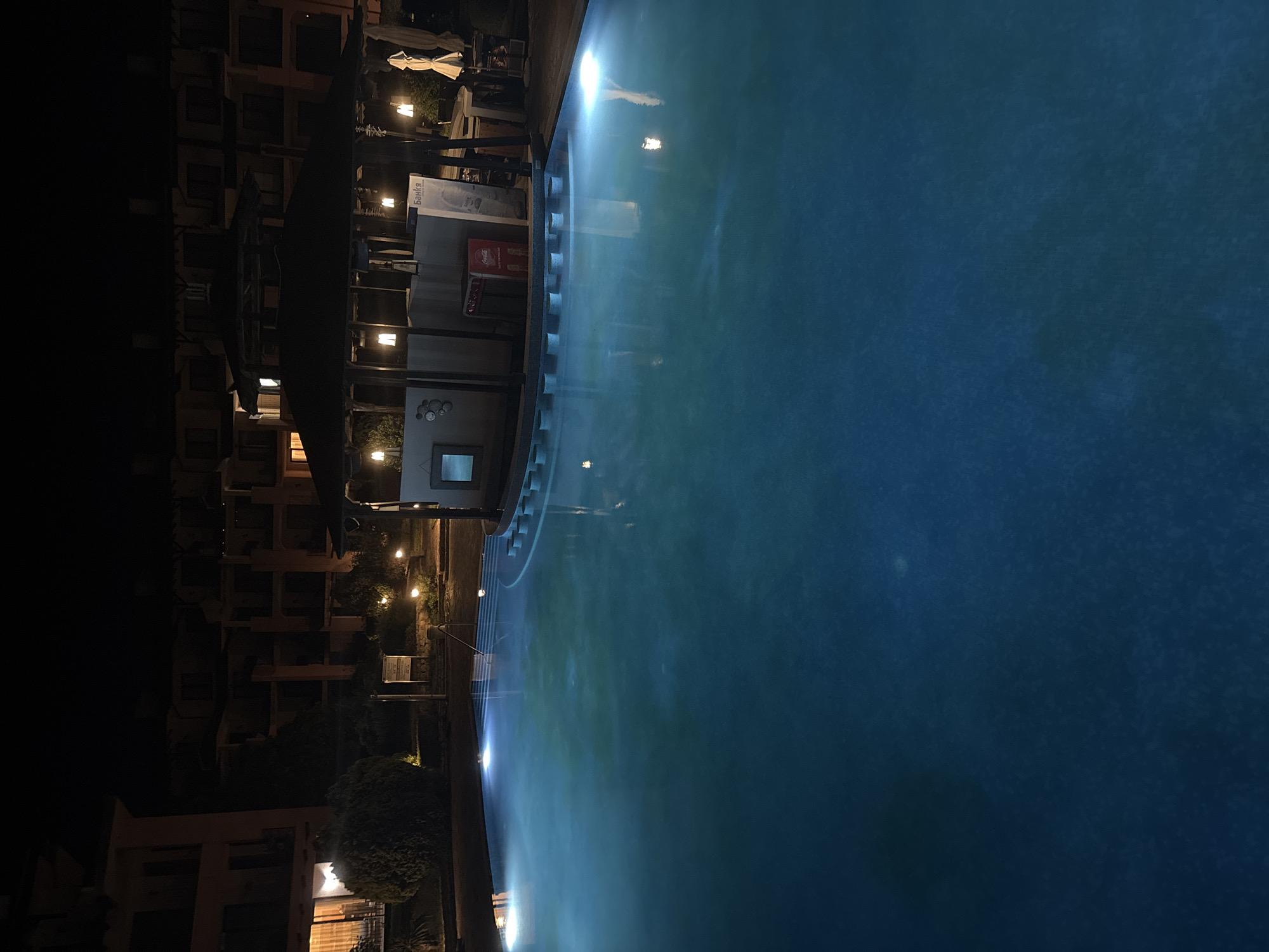 An outdoor swimming pool at night in Topola, Bulgaria, is illuminated by underwater lights, casting a blue glow on the water. In the background, a building with multiple balconies and windows is visible, along with a covered outdoor bar area with hanging lights. A Coca-Cola branded cooler is visible at the bar. The scene is peaceful and serene.