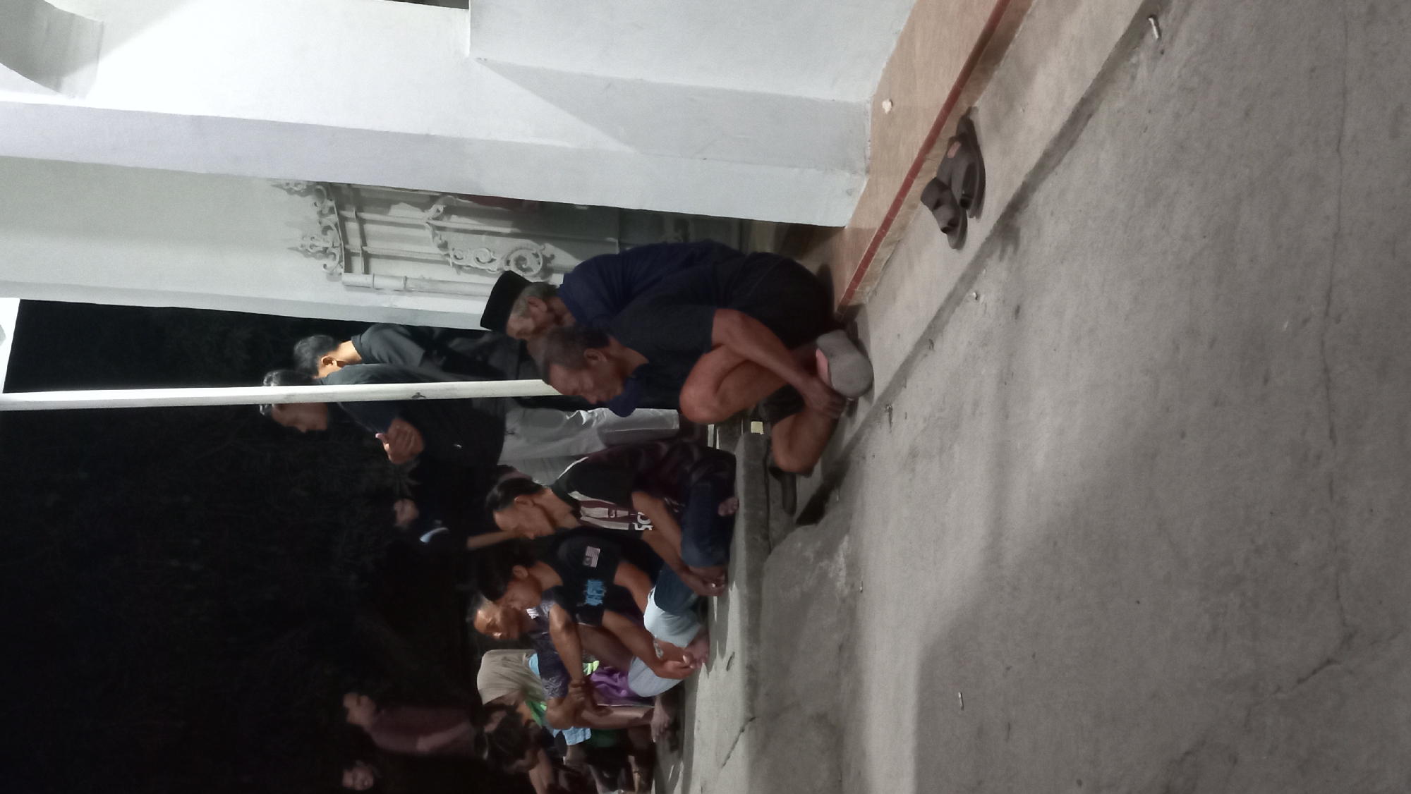 In Kedungmegarih, Indonesia, a group of men are gathered outdoors at night, sitting on the ground and on concrete steps. The scene appears to be a public space, possibly near a building with white pillars and decorative metalwork visible. The men are casually dressed, with some wearing t-shirts and others a more traditional cap. Some are looking in the same direction, suggesting they are observing something or participating in a shared activity. The lighting is dim, indicating it's nighttime, and the overall atmosphere is calm.