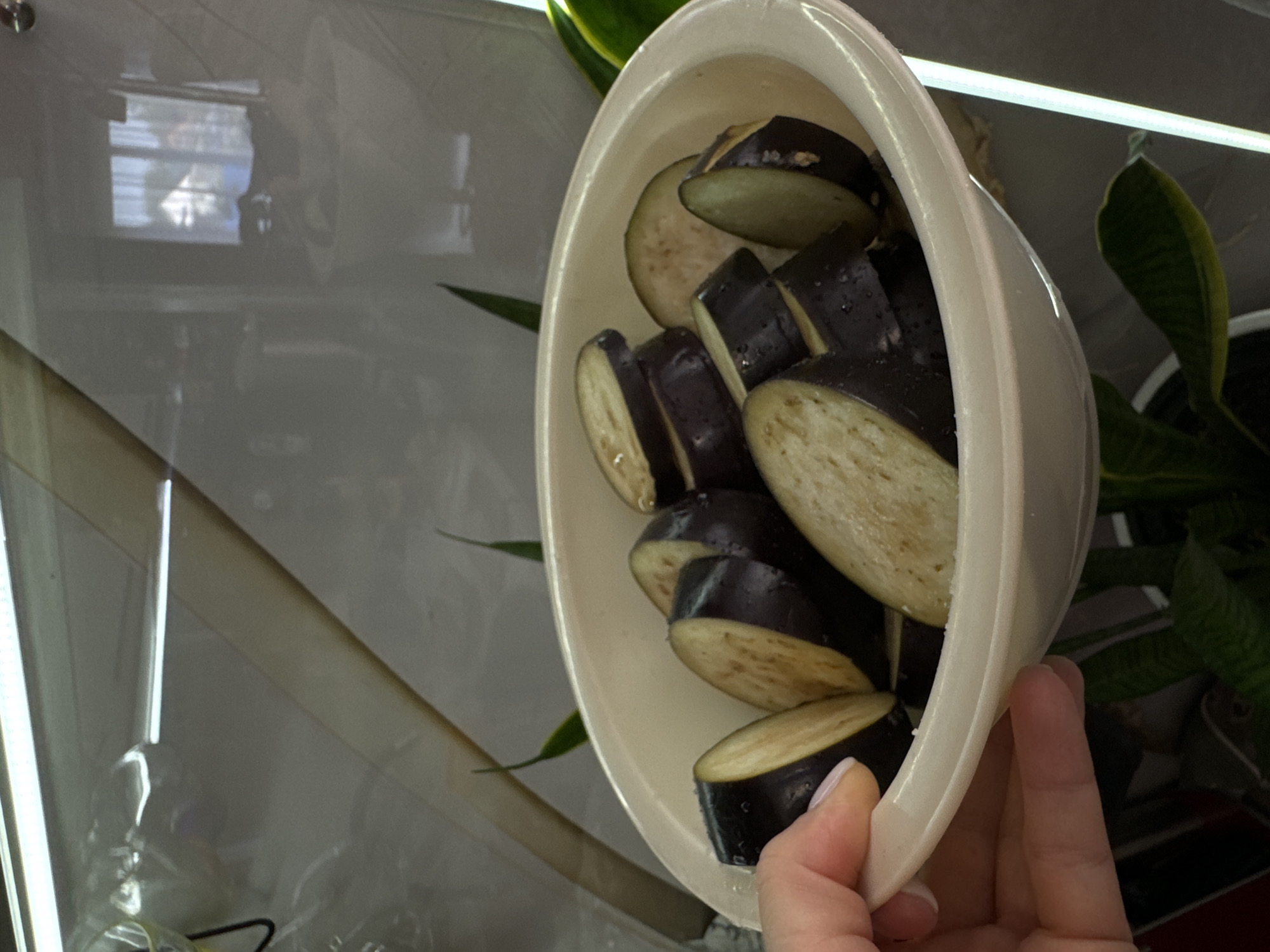 In a domestic kitchen environment in Ponazyrevo, Russia, a person's hand is visible holding a white bowl filled with sliced eggplant. The eggplant slices are dark purple on the outside and have a pale, creamy interior, some showing light brown speckling. The bowl appears to be made of plastic or a similar material and has a slightly raised rim. The background is out of focus, showing hints of kitchen cabinetry and possibly a window with curtains, suggesting an indoor setting. Green leaves of a houseplant are visible in the lower portion of the frame.