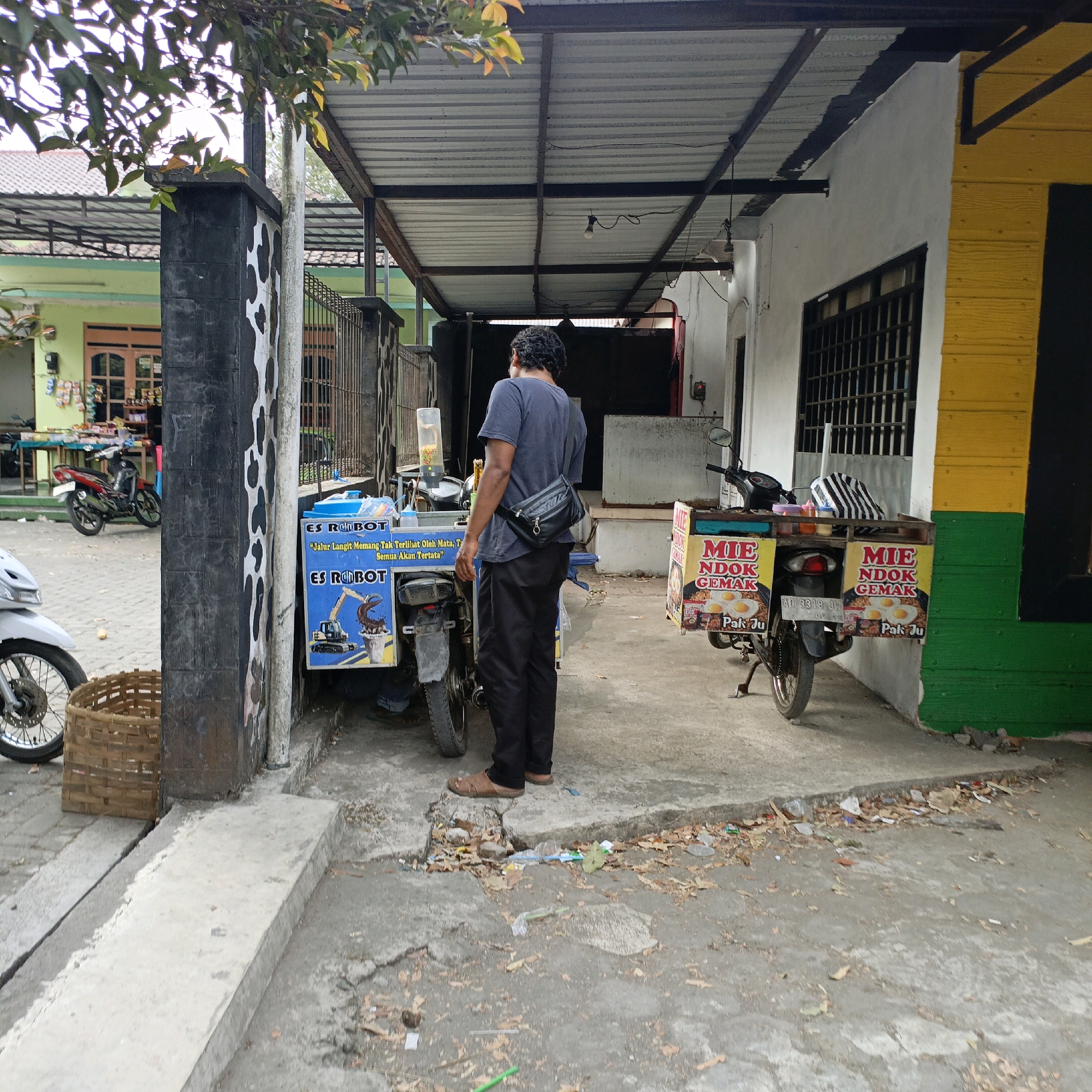 The image depicts a street scene in Salatiga, Indonesia, likely in front of a commercial establishment. A man, wearing a dark blue t-shirt and black pants, stands facing away from the camera, his back to two food carts mounted on motorcycles. The food carts are decorated with colorful signage. One cart, blue and white, displays images of ice cream and text in Indonesian, including "ES ROBOT". The other cart, yellow and red, advertises "MIE NDOK GEMAK" with images of fried eggs. A white motorcycle is partially visible on the left, with a woven basket next to it. In the background, a building with a yellow and green facade is visible, featuring a barred window. The ground is paved with concrete and shows signs of wear and litter. A tree with green and yellow leaves overhangs the scene from the upper left. The overall impression is a snapshot of daily life and small businesses operating on a street in Salatiga.