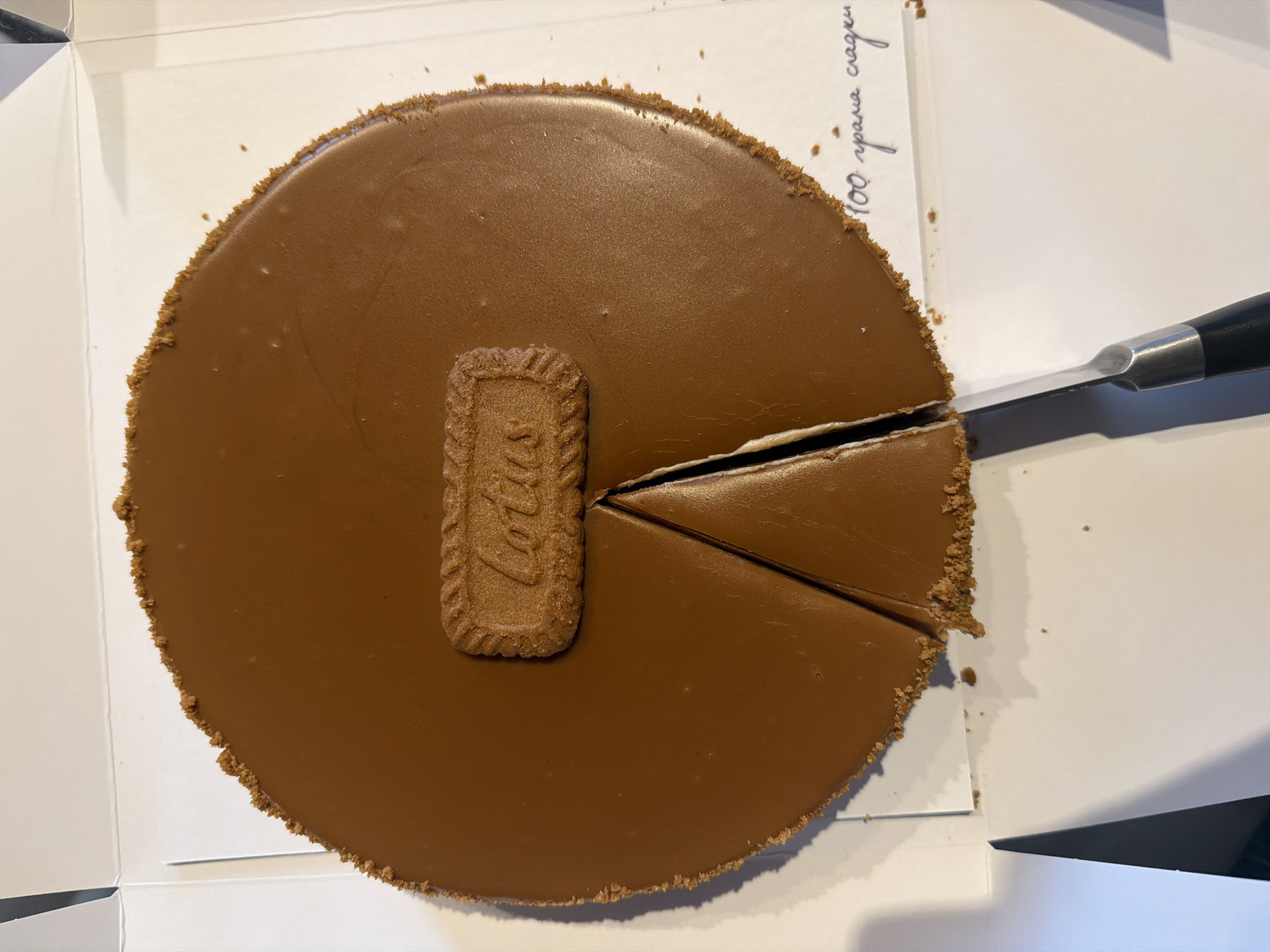 The image shows a close-up, overhead view of a round, chocolate-frosted cake with a cookie on top, resting in a white box. A slice has been cut from the cake, and a knife with a black handle is positioned to cut another slice. The cake has a crumbly, biscuit-like crust around its edge. To the right of the cake, handwritten text in Cyrillic script on the white surface reads "100 грама сладки" which translates to "100 grams of sweets". The scene appears to be indoors, possibly in a kitchen or dining area, given the presence of the cake and serving implements. The lighting suggests it is daytime. There are no people visible in the image. The overall impression is that of a dessert ready to be served.