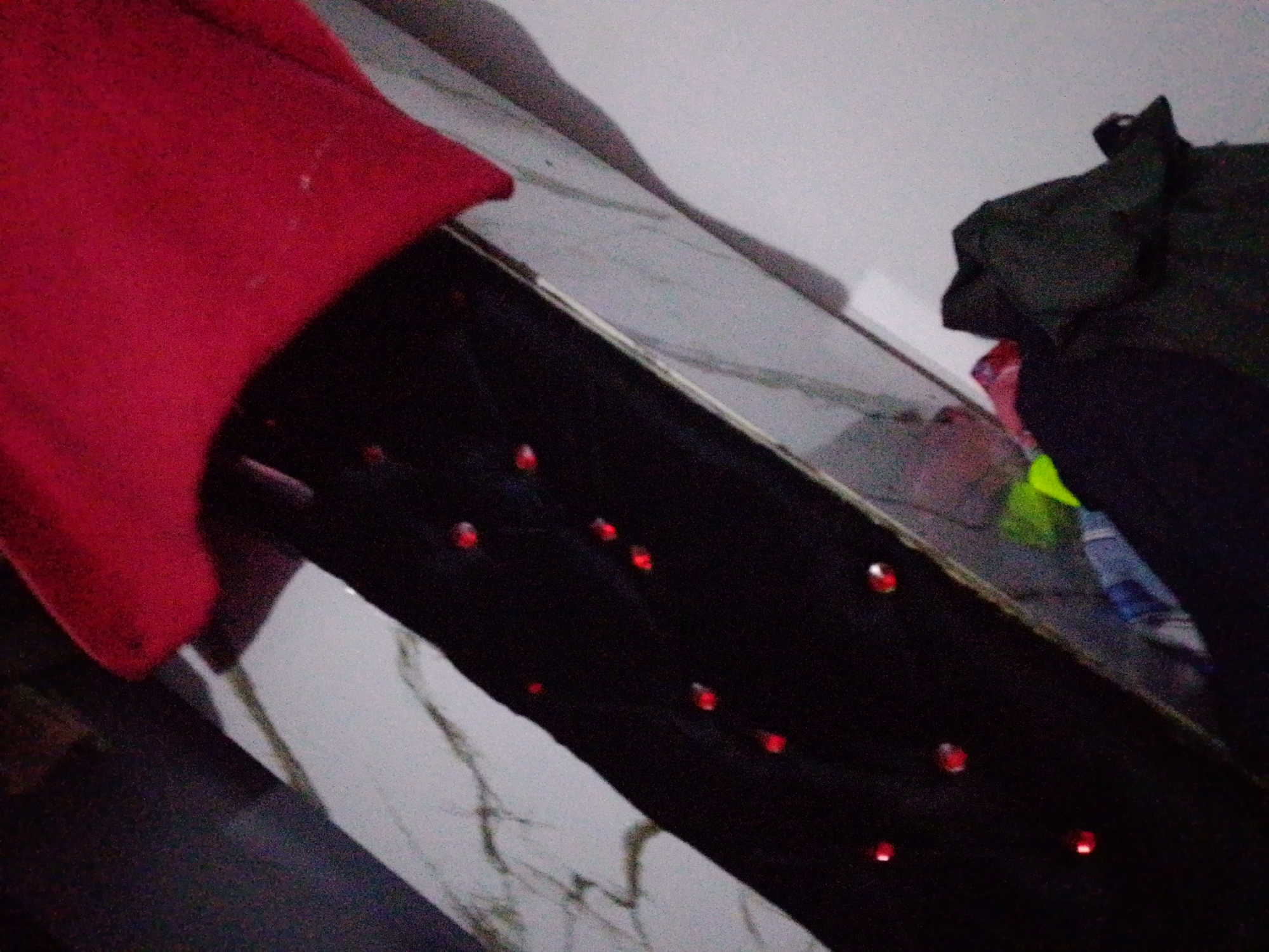 The image depicts a close-up view of a dark fabric, possibly a dress or scarf, adorned with small, bright red embellishments that appear to be sequins or beads. These are arranged in a scattered pattern across the fabric. In the background, a large portion of a red garment is visible, suggesting it is draped over or near the dark fabric. To the right, a dark, possibly green or black, piece of clothing is seen, with hints of other colorful items like a green and blue striped cloth and a pink item peeking out from underneath. The setting appears to be indoors, with a wall and a reflective surface, possibly a table or floor with a marble-like pattern, visible in the background. The lighting is dim, creating a somewhat blurry and shadowed effect. There are no people or clear indications of the specific location within Jalingo, Nigeria, other than what is provided in the prompt.