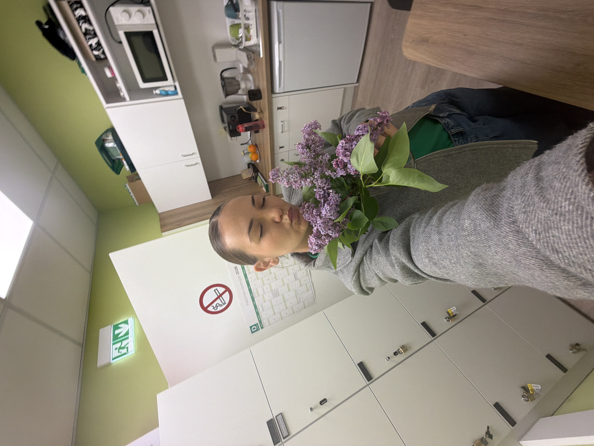 A selfie shows a person holding a bouquet of lilac flowers. The person is wearing a grey sweater and jeans. In the background are lockers, a "no smoking" sign, and an emergency exit sign. Behind the person are a refrigerator, cabinets, and a microwave. The location is most likely indoors. The city of Sofia, Bulgaria, is not discernible from the image.