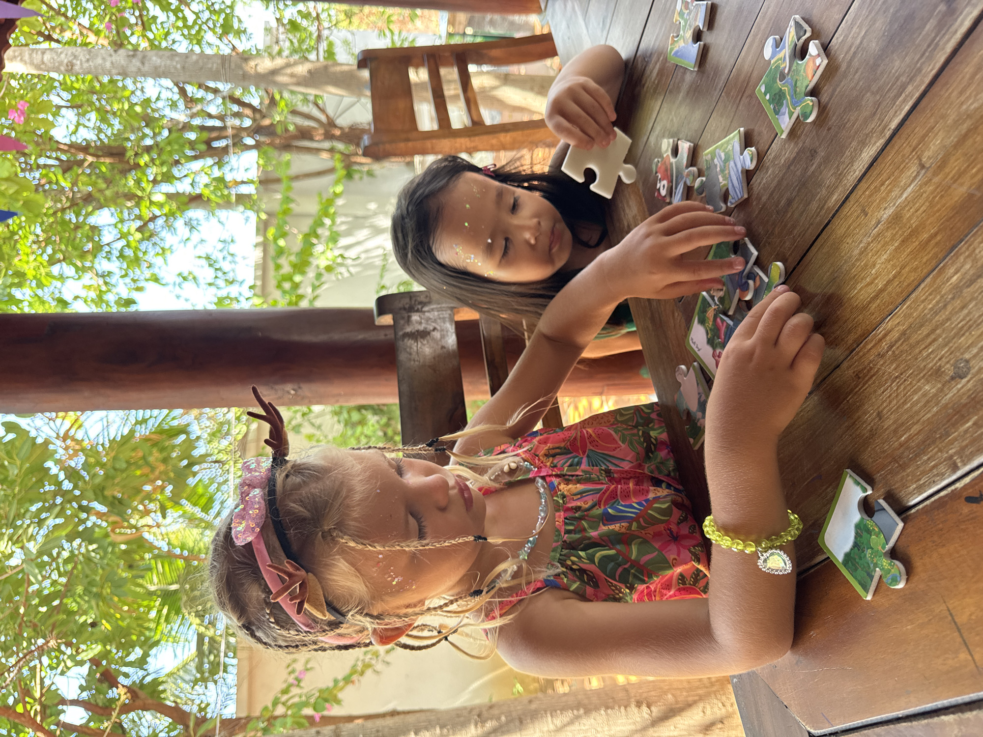 Two young girls are sitting at a wooden table, working on a jigsaw puzzle. The girl closer to the viewer has blonde hair styled with braids, a floral headband with antler-like protrusions, glitter on her face, and a bright, floral-patterned dress. She is wearing a beaded bracelet with a heart-shaped charm. The other girl has dark hair and glitter on her face, and she is holding a white puzzle piece. Several other puzzle pieces are scattered on the table. The scene takes place outdoors, under a wooden structure, with trees visible in the background. The exact location within Mũi Né, Vietnam, cannot be determined from the image.