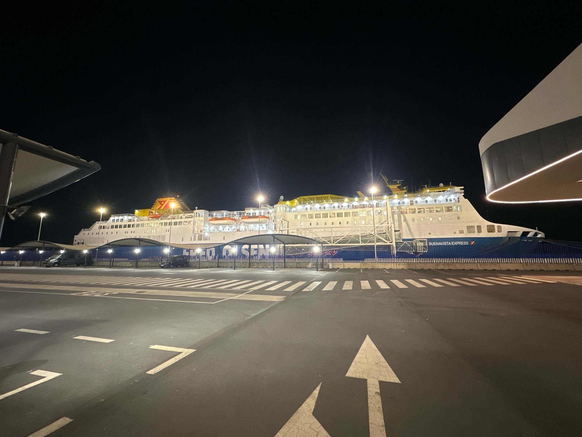 The image depicts the "BUENAVISTA EXPRESS" ferry docked at the port of Palos de la Frontera, Spain, at night. The ferry, a large white vessel with blue accents and yellow upper decks, is illuminated by onboard lights and external floodlights. The ferry has several lifeboats visible on its side. In the foreground, a paved area with white road markings, including arrows and lane dividers, suggests a parking or waiting area for vehicles. Several vehicles, including vans and cars, are parked under covered shelters behind a blue fence in front of the ferry. A modern, curved building with a glowing light strip is visible to the right of the frame. The sky is dark, indicating nighttime.