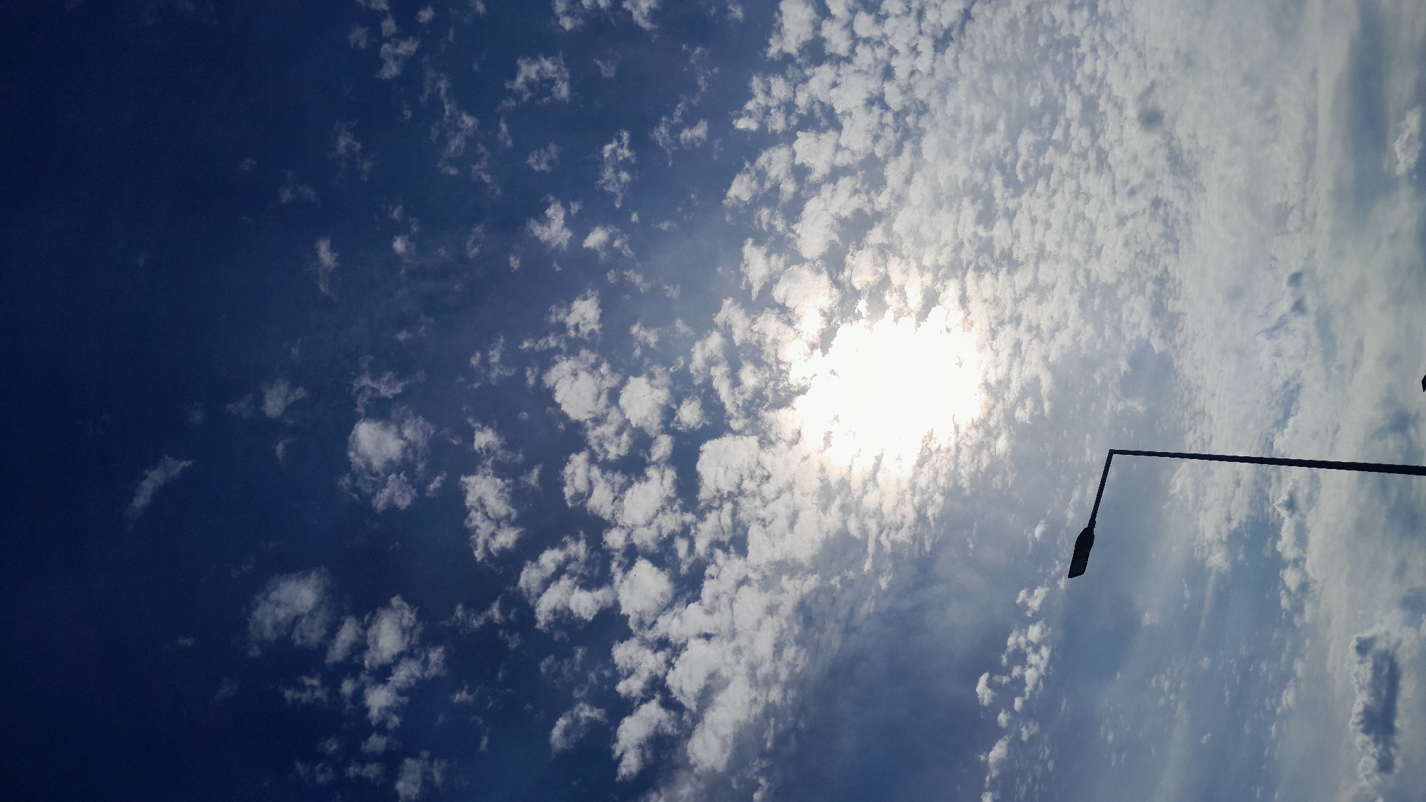The image depicts a clear blue sky filled with scattered white cumulus clouds. The sun is bright and partially obscured by the clouds, creating a luminous effect in the upper center of the frame. A single black lamppost stands in silhouette against the sky on the left side of the image. The lamppost has a tall, slender pole topped with a rectangular light fixture. The bottom of the image shows a sliver of dark ground. The image does not contain any people or specific identifiable landmarks to determine the location within Krakow, Poland. The scene appears to be an outdoor urban environment, likely during daylight hours.