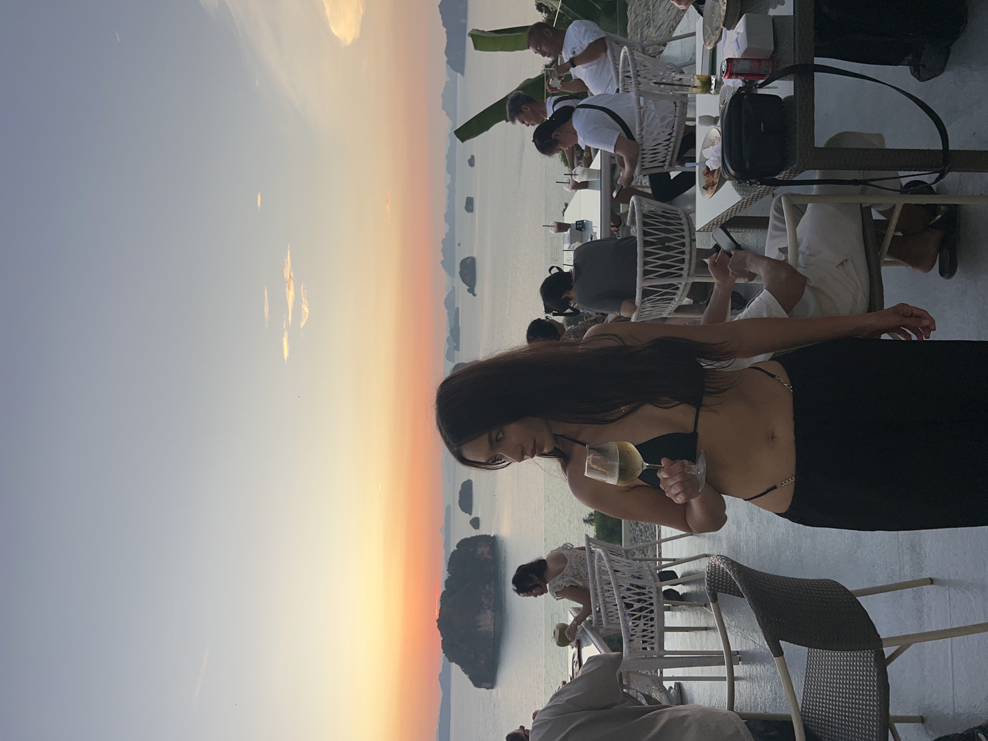 Here is a description of the image:
The image depicts a sunset scene in Khao Thong, Thailand. In the foreground, a woman with long dark hair is holding a glass of white wine. She is wearing a black outfit with straps. Behind her, there are tables and chairs with other people dining. In the background, there's a view of the ocean with islands and the colorful sunset sky.