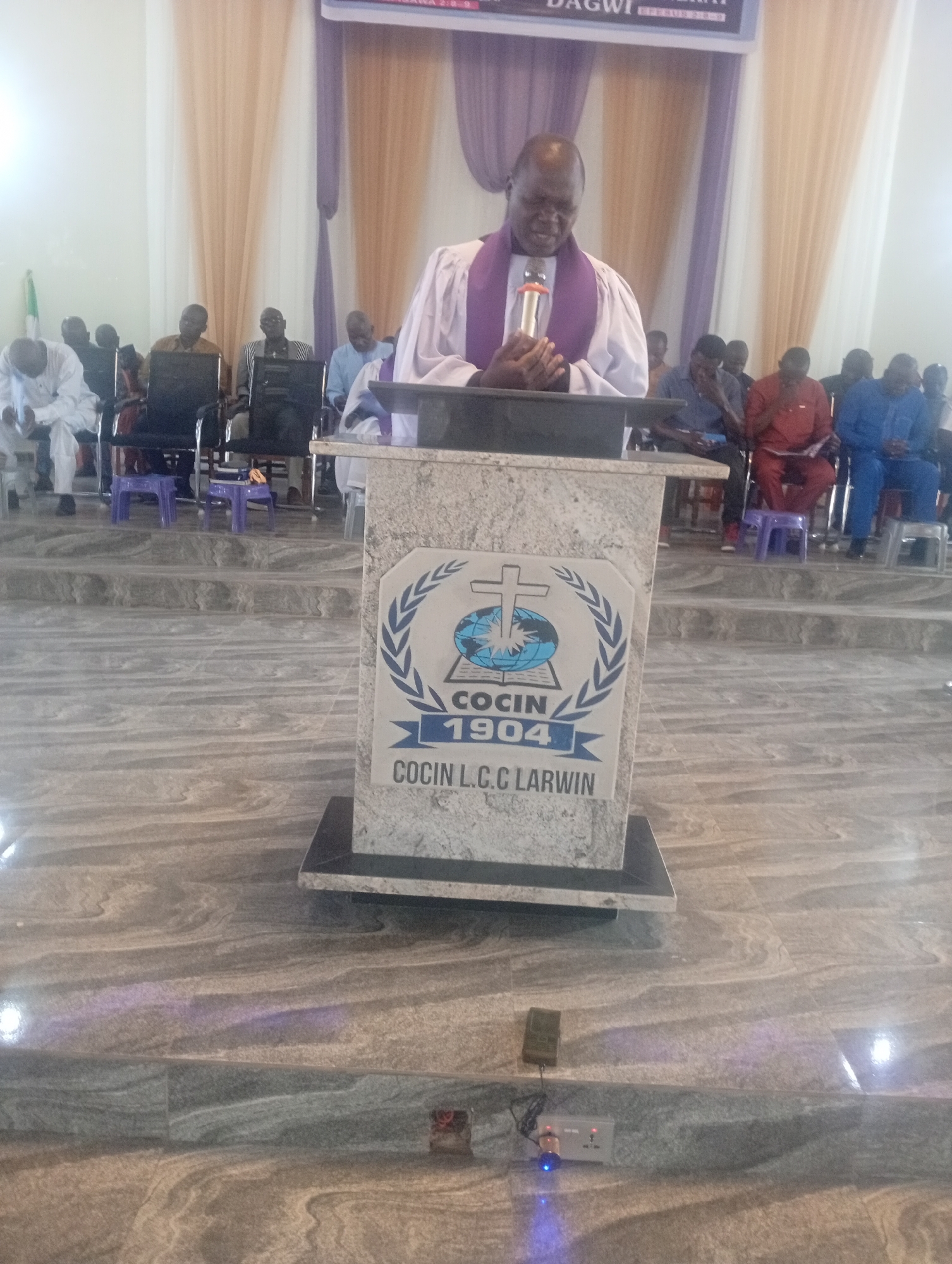 A medium shot captures a man, likely a preacher, standing behind a podium inside what appears to be a church in Unknown, Unknown. He is wearing a white robe with a purple stole and is speaking into a microphone. Behind him, a backdrop of peach and purple drapes hangs. Seated behind the podium are rows of men, presumably congregants. The podium bears the insignia "COCIN 1904 COCIN L.C.C LARWIN". The floor is tiled in grey with subtle patterns. In the foreground, an electrical outlet and a small device are visible on the floor.