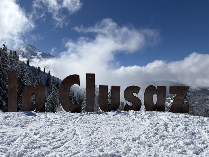 The image displays a panoramic view of a snow-covered mountainous landscape in La Clusaz, France, centered on large, rust-colored metal letters spelling "la Clusaz." The letters stand upright on a deep layer of disturbed snow in the foreground, showing numerous tracks from skis or footwear. Behind the prominent signage, a dense forest of snow-laden evergreen trees covers a steep mountain slope. Further in the background, a prominent snow-capped mountain peak is partially obscured by low-hanging white and grey clouds. The sky above is a clear, vibrant blue with scattered white clouds, and a faint white contrail is visible in the upper right. The scene is illuminated by natural daylight, indicating clear weather conditions. No individuals are present, nor are any active interactions depicted, though the snow tracks suggest recent human presence in a recreational context. The specific location is a scenic viewpoint within the La Clusaz ski resort area.