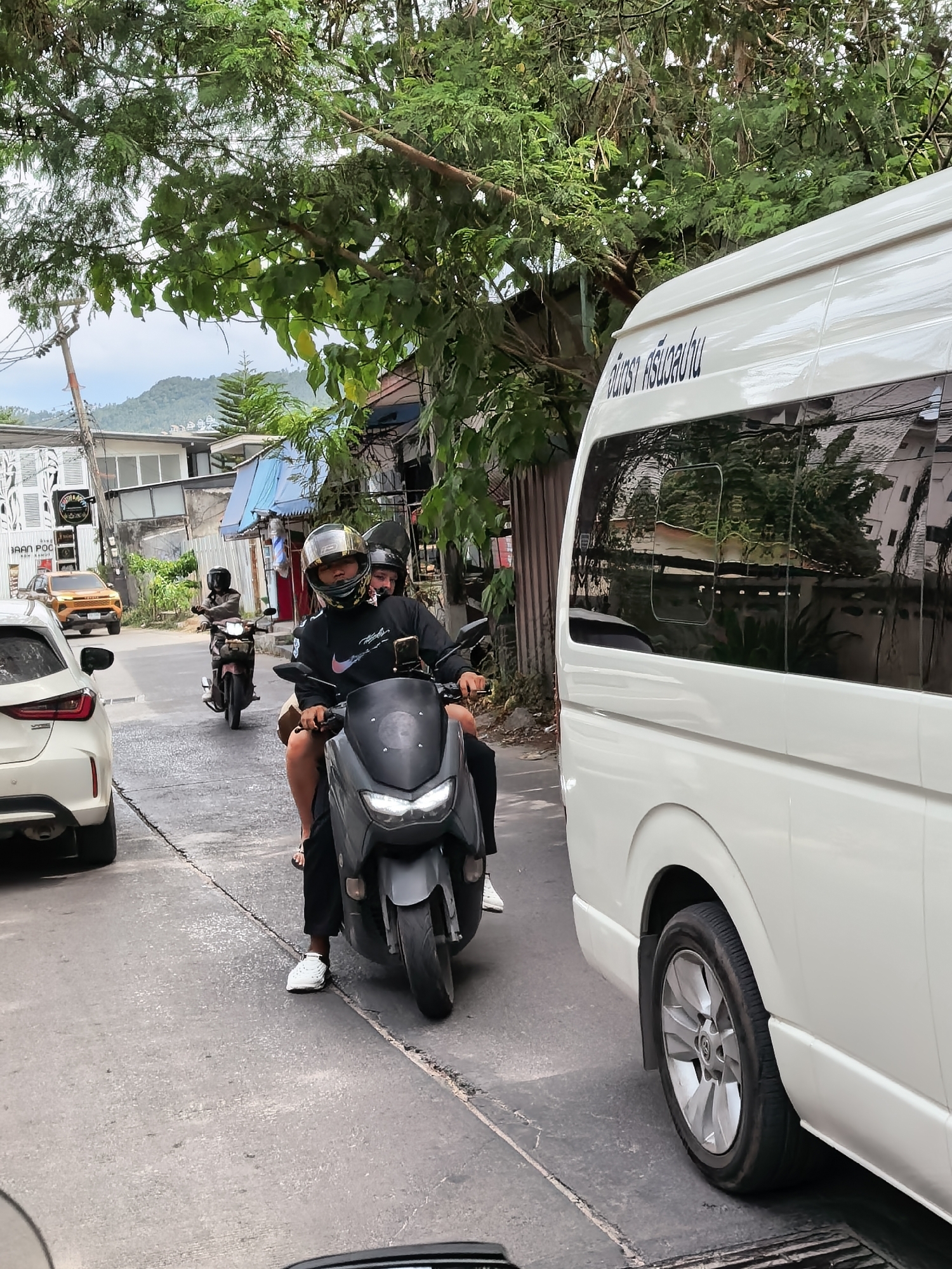 In the city of Ko Samui District, Thailand, a white van is parked on the right side of a street. On the left, a white car is also parked. Two individuals are riding a dark-colored scooter in the street. The person in front is wearing a black shirt, black pants, and white sneakers. They are also wearing a helmet. The person behind them is wearing a black shirt and is also wearing a helmet. The scooter is facing away from the camera. Further up the street, another scooter with a single rider wearing a helmet is visible. Buildings and trees line the street. The sky is overcast.