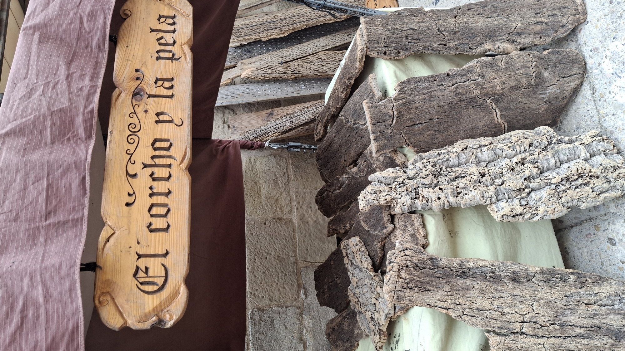 The image depicts a stall at an outdoor market in La Fresneda, Spain. A wooden sign with ornate carving reads "El corcho y la pela." In the foreground, various pieces of rough, textured bark, likely cork, are displayed on a light-colored cloth. Behind the bark, a stone wall forms the backdrop. To the right, several long, weathered wooden planks, some with visible perforations, are leaning against a stall structure. The overall scene suggests a rustic market setting selling natural materials.