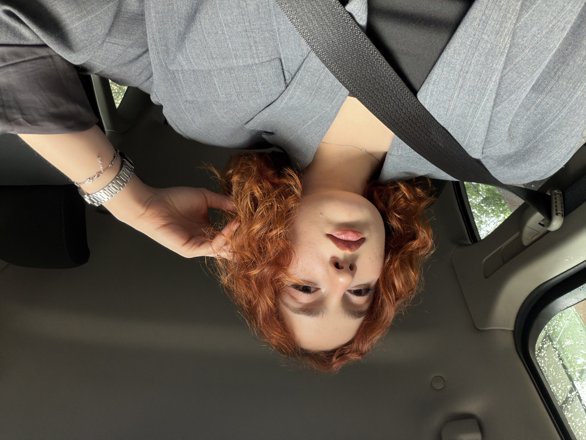 A red-haired woman in a gray pinstripe suit is wearing a seatbelt in a car. She is holding her hair with her left hand, which is adorned with a bracelet and a watch. It is raining outside, and the droplets are visible on the car window. The location cannot be determined from the provided image.