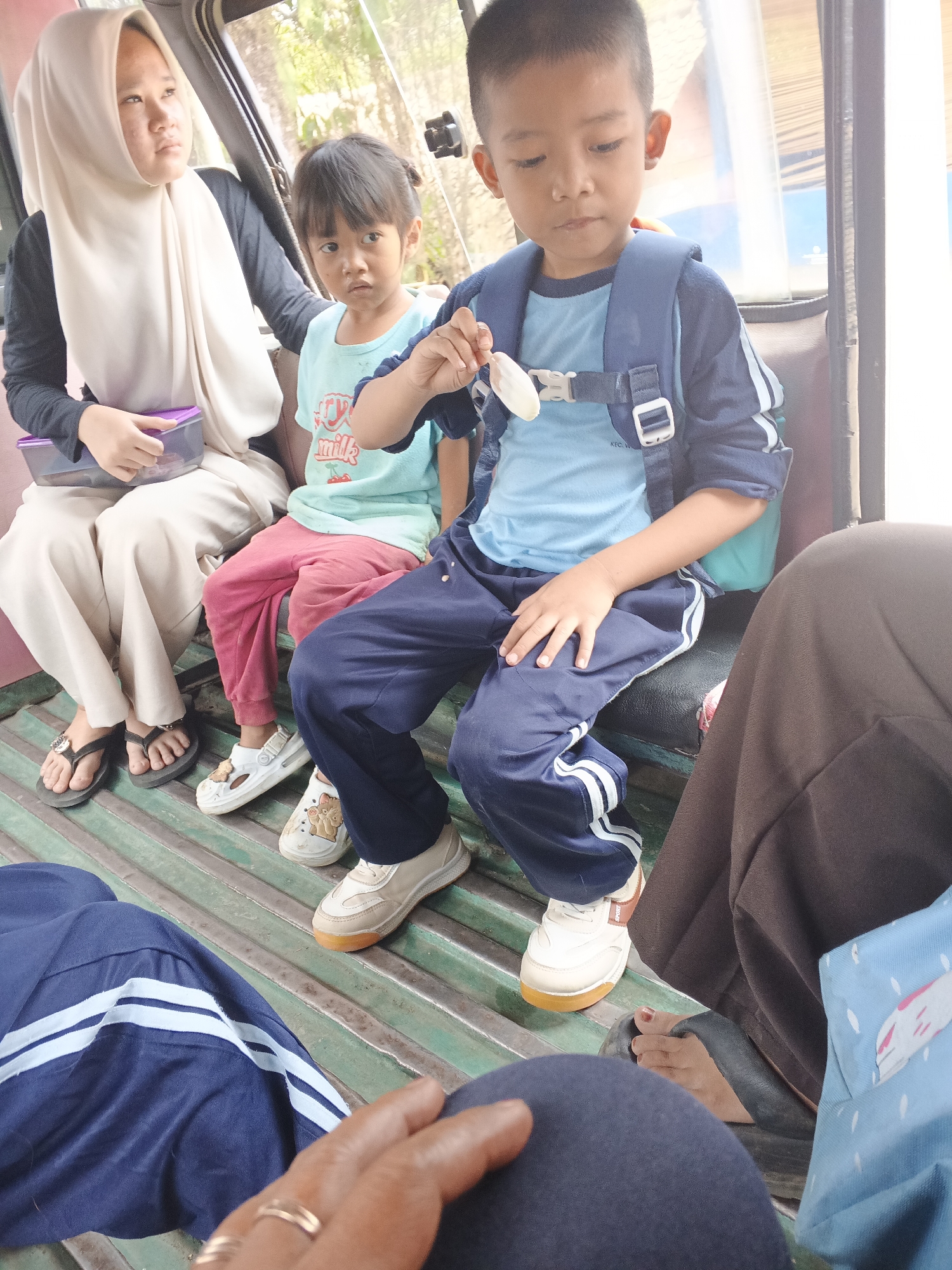 In Warungkiara, Indonesia, four people are seated inside a vehicle, likely a van, on a ribbed green metal bench. The person on the far left, wearing a cream-colored hijab and dark long-sleeved shirt, holds a purple container. Next to them, a young child in a light blue t-shirt with a "milk" graphic and pink pants sits with their legs crossed, looking forward. To their right, a young boy with a blue backpack and a blue long-sleeved shirt eats from a light-colored food item. His right hand is raised to his mouth. The fourth person, seated on the far right, is partially visible, wearing brown pants. The floor of the vehicle is also made of ribbed green metal. The background shows trees and sunlight, indicating an outdoor environment.