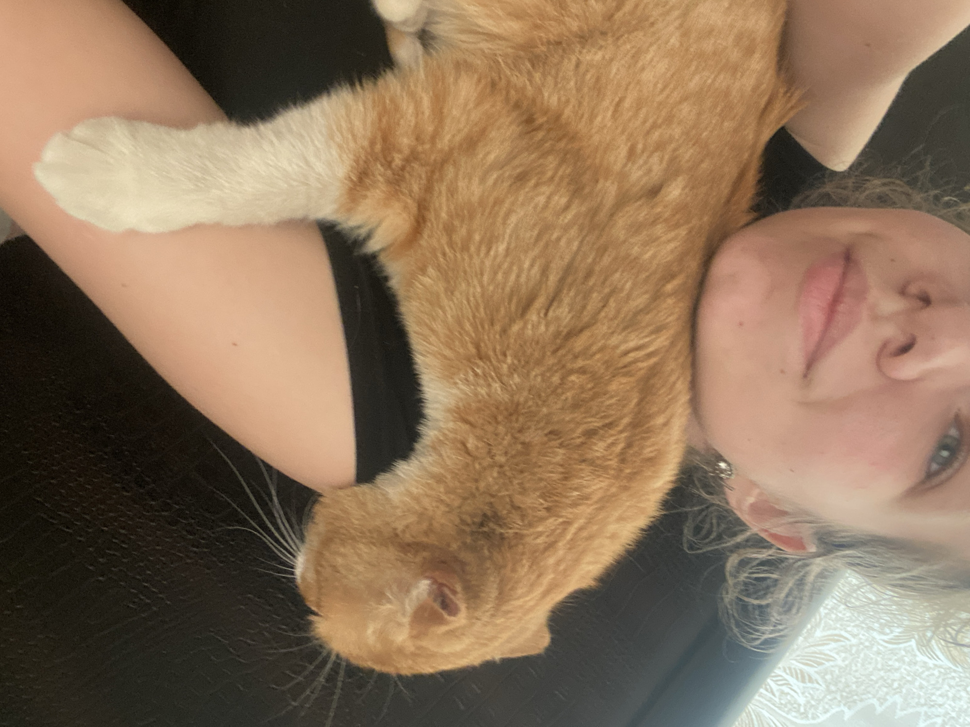 A woman is lying on her back, holding an orange cat. The cat is lying lengthwise on the woman's chest and arm. The cat has white paws. The woman is looking at the camera. The location and city, Pakhynia, Ukraine cannot be determined.