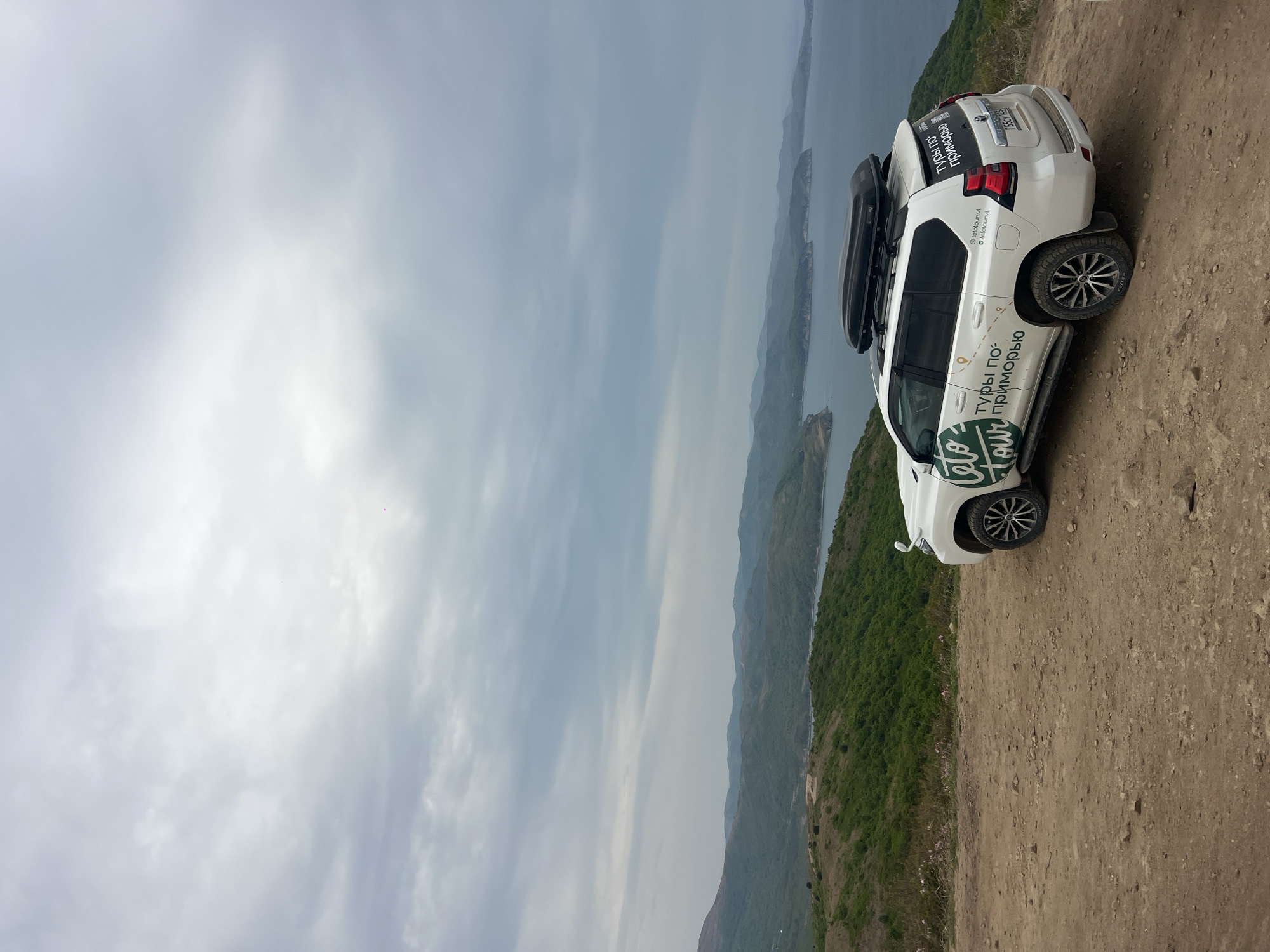 A white SUV with a roof rack is parked on a dirt ledge overlooking a body of water with mountainous terrain in the distance under an overcast sky. The SUV has Russian lettering on its side and rear. The SUV's license plate is partially visible. The location is Vityaz', Russia. The image appears to be taken from slightly below the vehicle, angled upwards.