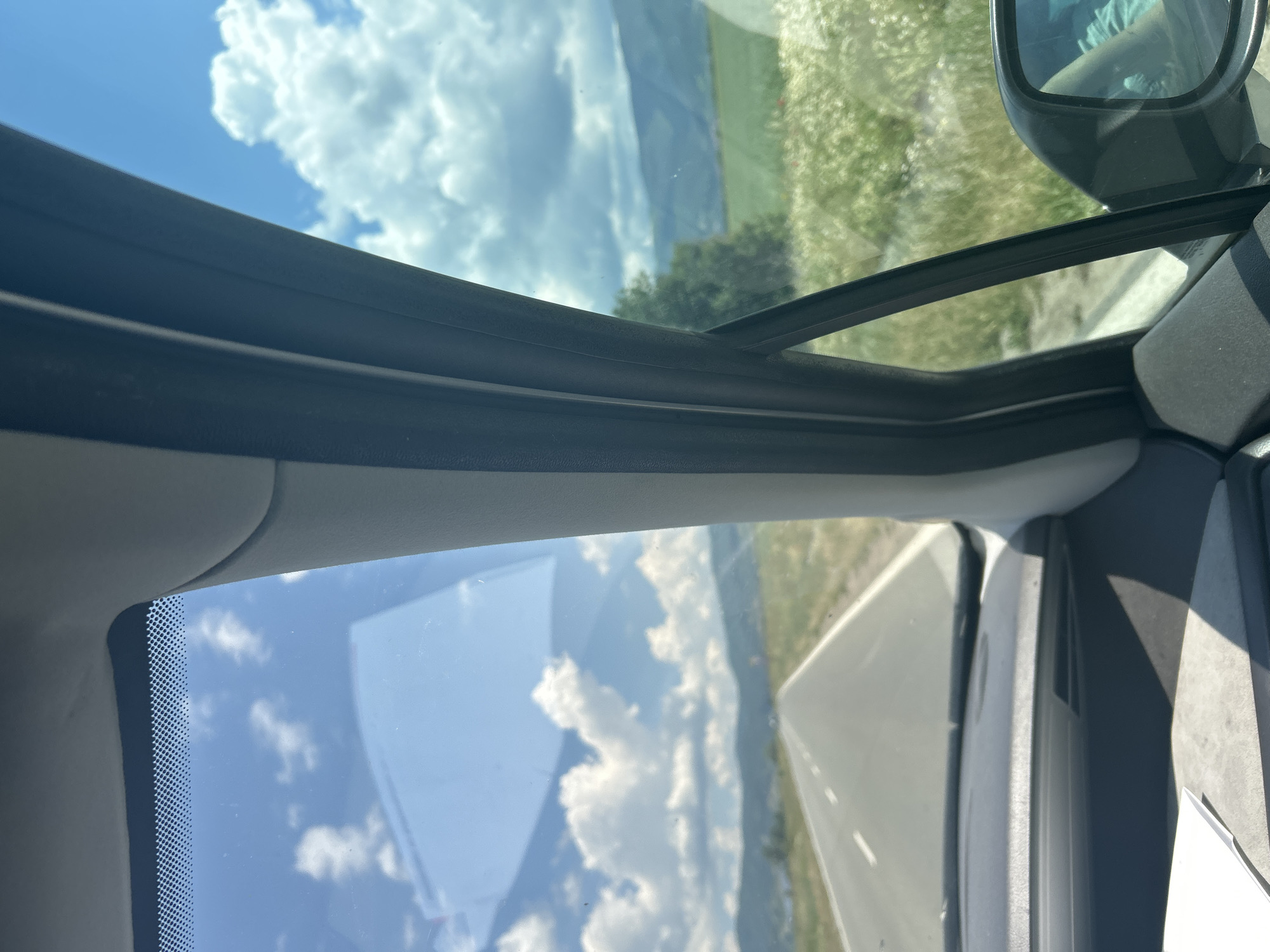 The image, taken from inside a vehicle, captures a view through the windshield and side window. A highway stretches into the distance, flanked by green fields and distant mountains under a bright blue sky dotted with white clouds. The car's interior, including the dashboard and parts of the A-pillar, are visible in the foreground. The scene depicts a journey through the Bulgarian countryside, potentially near the village of Yahinovo, known for its natural beauty and proximity to mountainous terrain.