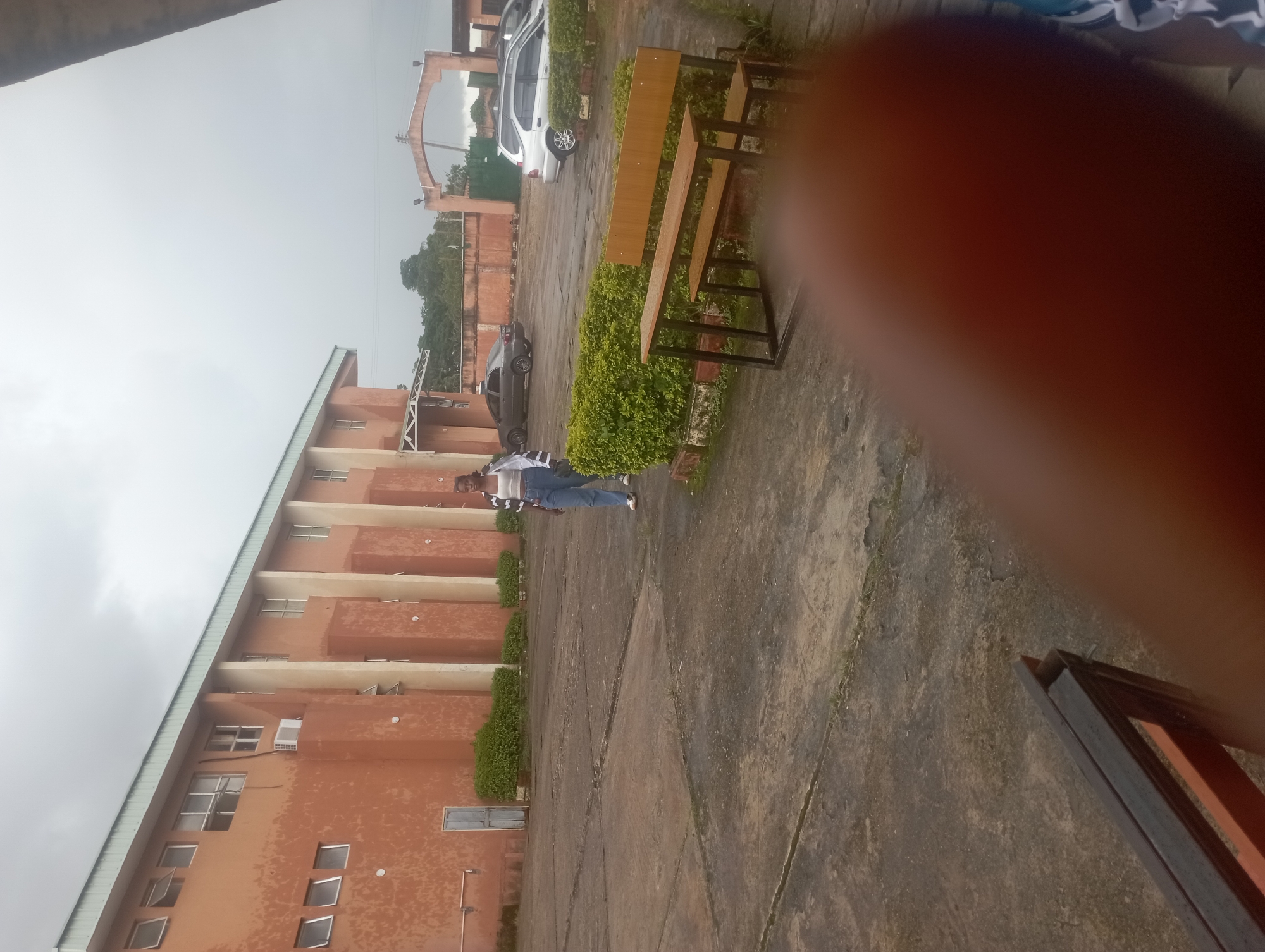 In Jos, Nigeria, a young woman walks across a paved courtyard in front of an orange building. The building features numerous windows, some open, and vertical beige pillars. A blue door is visible on the left side of the building, and a metal bench sits in the foreground. Further back, several cars are parked near a large archway. The sky is overcast, suggesting a cloudy day. The scene appears to be an outdoor area adjacent to an educational or institutional building.