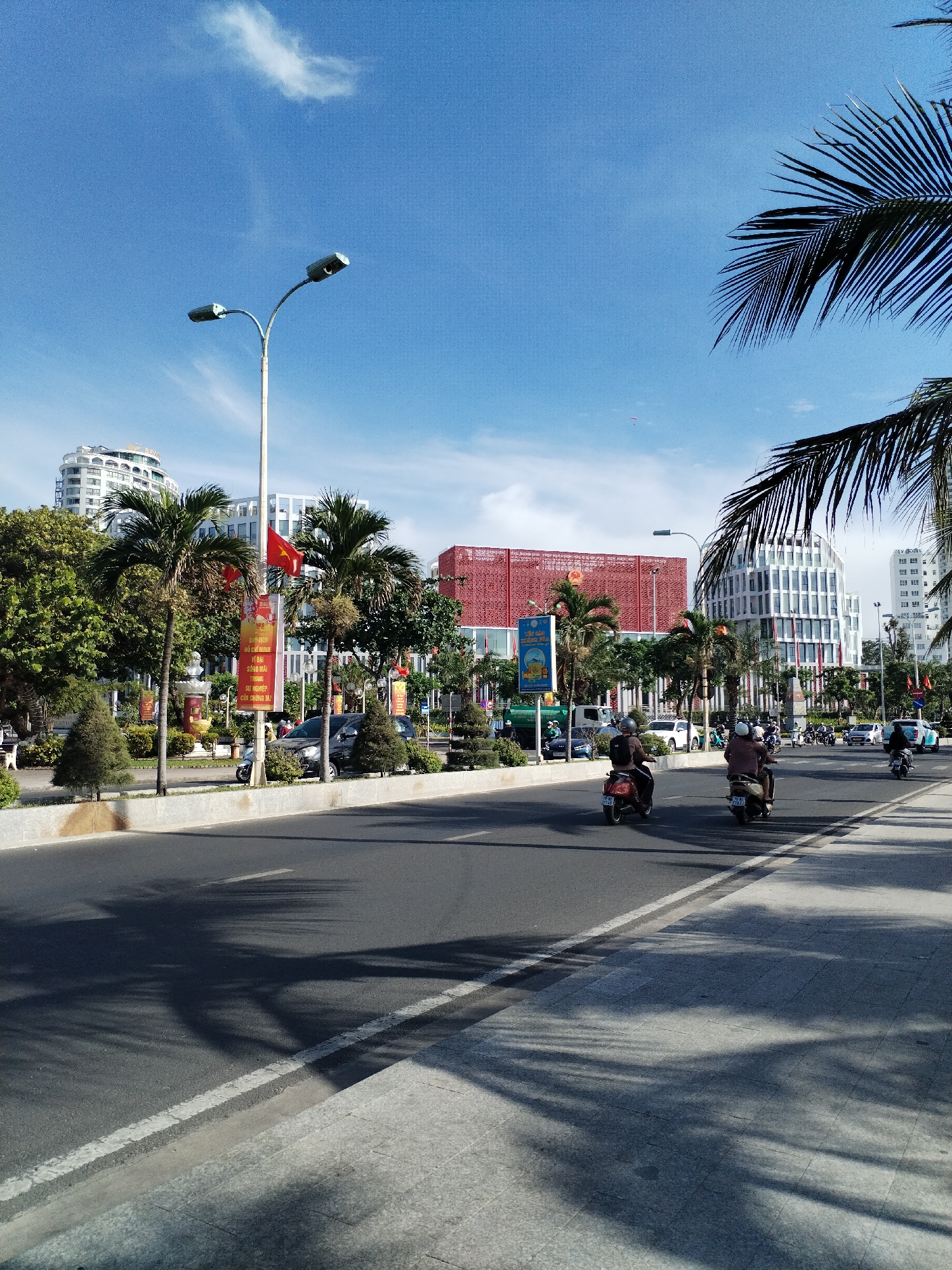 The image depicts a street scene in Nha Trang Ward, Vietnam, characterized by a wide asphalt road with motorcycles in motion. The road is flanked by sidewalks with patterned shadows from palm trees. The street is bisected by a planted median with palm trees, bushes, and decorative flags.
In the background, various architectural structures are visible, including modern high-rise buildings, one with a red lattice facade and another with a curved, white facade. Utility poles with lights are present along the roadside. The sky is predominantly blue with sparse white clouds.