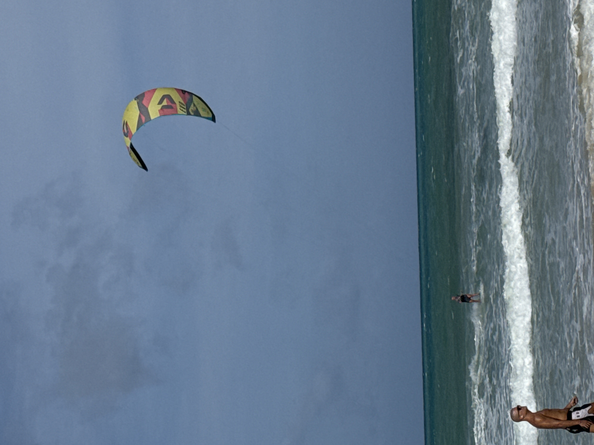 A man walks on a beach in Rawai, Thailand, as a kitesurfer rides in the ocean in the background. The kitesurfer is positioned in the middle of the frame, riding a white board and being pulled by a colorful kite. The man in the foreground is walking towards the right of the frame, wearing sunglasses and black and white swim trunks. The ocean is blue-green with white waves, and the sky is a muted blue with some clouds.