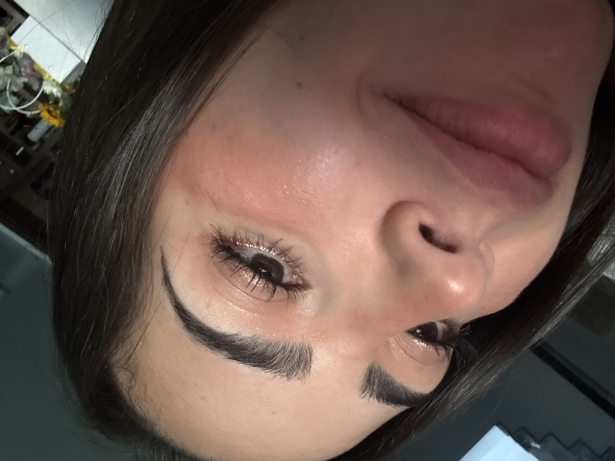 A close-up, upward-angled portrait of an individual, likely female, taken indoors within San Bernardino Tlaxcalancingo, Mexico. The subject's dark hair is visible along the top and left edges of the frame. The right eye is prominent, featuring dark, sculpted eyebrows, extended eyelashes, and a subtle shimmer of light-toned eyeshadow on the eyelid. The iris is dark. The right cheek exhibits a light reddish hue, consistent with blush or natural flush. The nose is partially visible, and the lips are slightly parted, displaying a natural pink tone and forming a faint smile. The subject's skin has a visible sheen. The background is dark and out of focus, revealing indistinct objects in the upper right, including a white, structural element and a blurred arrangement with yellow floral elements.