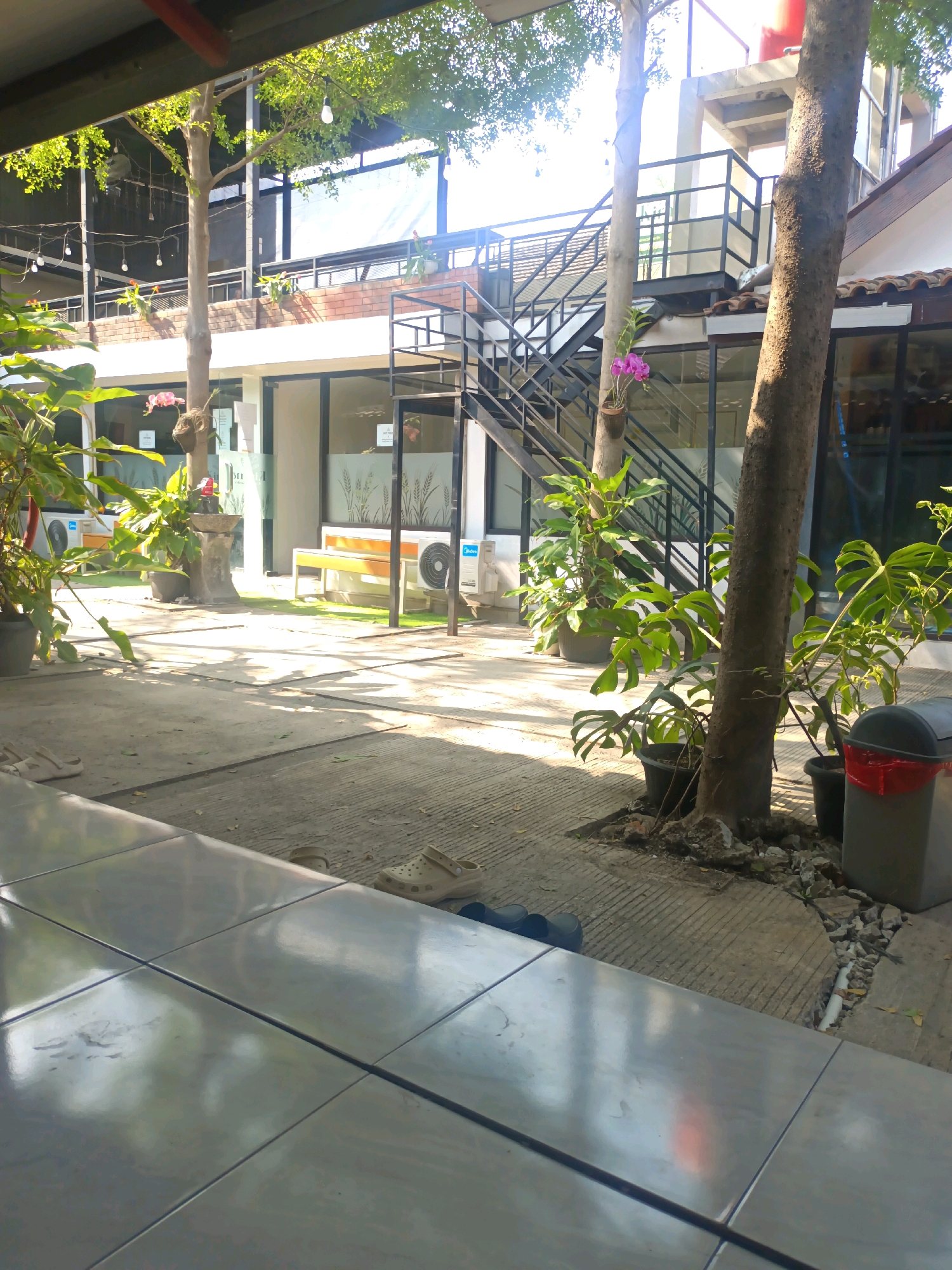 The image depicts an outdoor courtyard area, possibly at a cafe or restaurant, in Kerangkulon, Indonesia. The foreground features a tiled surface, reflecting light. Beyond this, a paved pathway leads into the scene, bordered by trees and potted plants. In the middle ground, there is a white building with large windows and an outdoor staircase leading to an upper level. A yellow bench is visible near the building. The background shows more greenery and structures, with the sky partially visible through the trees. Sunlight creates strong shadows across the ground. There are no people visible in the image.