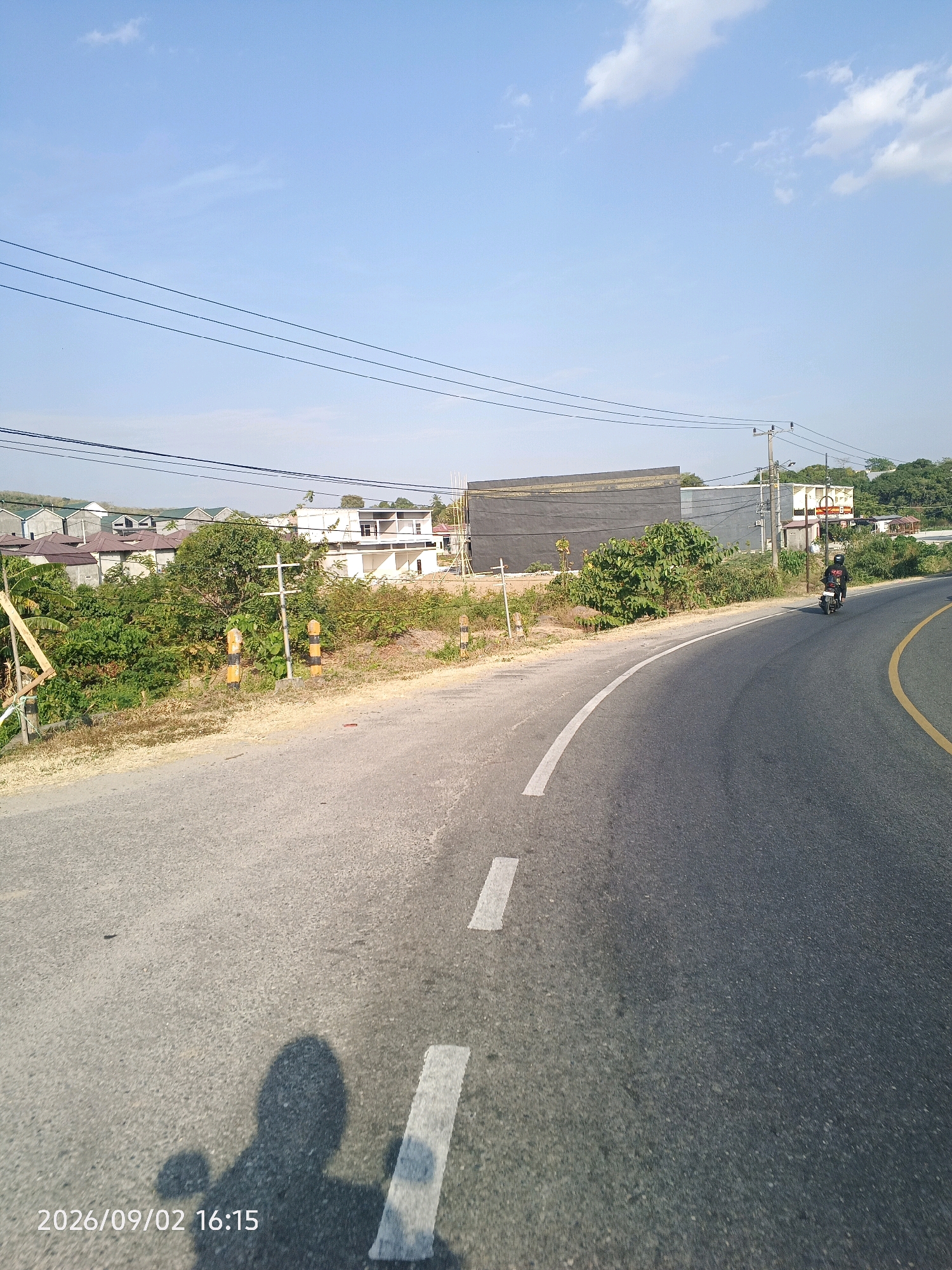 The image captures a roadside view of a developing area in Pinrang, Indonesia. A paved road curves to the right, marked with a yellow line on the right side and a dashed white line on the left. A single motorcycle rider, wearing black clothing and a helmet, is visible on the road in the distance. The left side of the road features a dry, dirt shoulder with sparse vegetation. Beyond the shoulder, there is a dense line of green bushes and trees, with several unfinished houses and buildings under construction in the background. Some of these structures have visible roofs and white walls. Overhead, power lines stretch across the frame against a clear blue sky with a few wispy clouds. The lower-left corner of the image displays a timestamp: "2026/09/02 16:15", and a shadow of what appears to be the photographer or their vehicle is cast on the road.