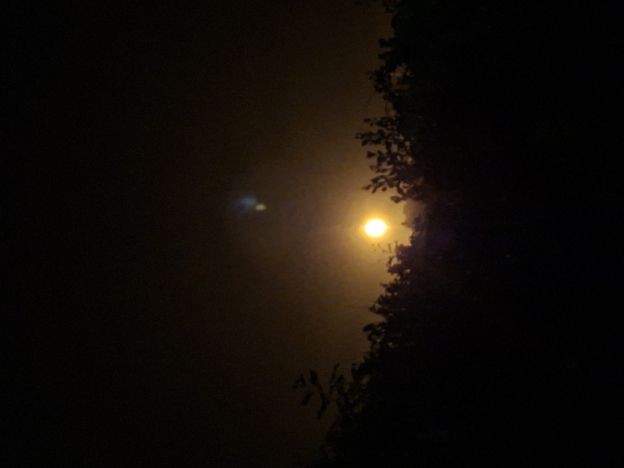A nighttime scene shows a bright, circular light source, likely a street lamp, partially obscured by silhouetted foliage, possibly a tree. The illumination creates a hazy, diffused glow in the surrounding area. The location is the city of Lhokseumawe, Indonesia.