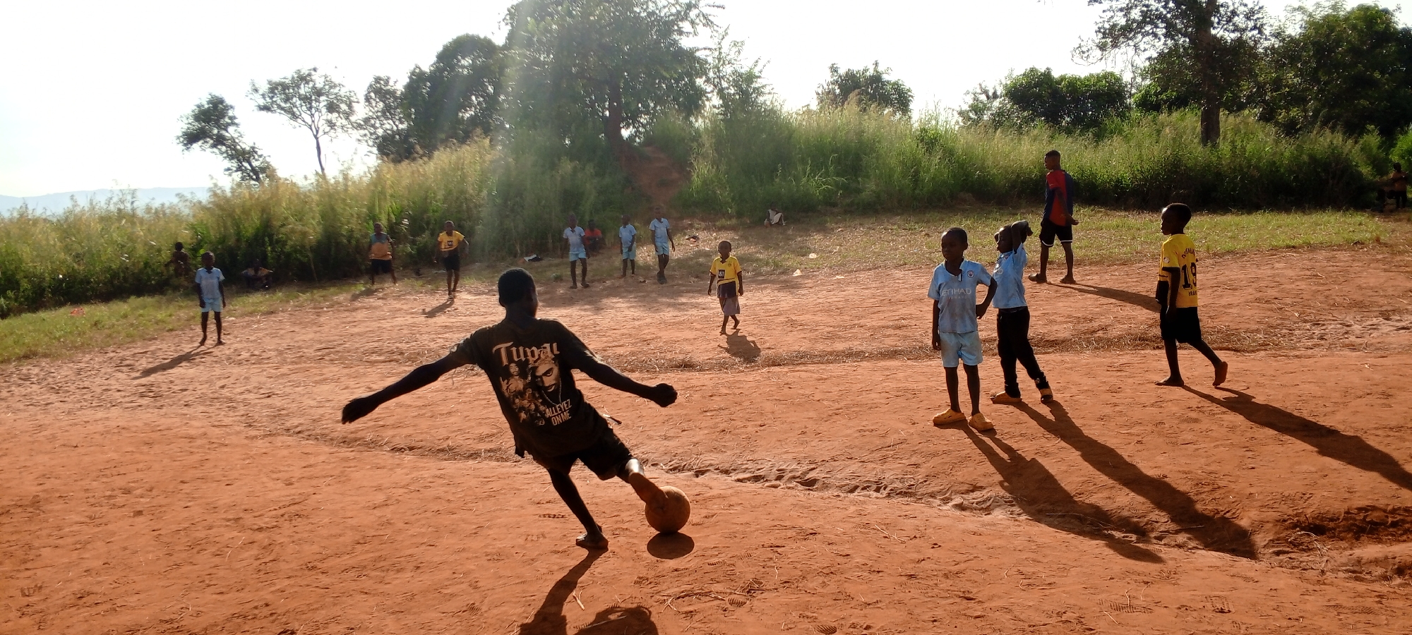 In an outdoor setting in Ngalula, Democratic Republic of the Congo, a group of children are playing soccer on a reddish-brown dirt field. The focal point of the image is a young boy in the foreground, wearing a dark T-shirt with an image of Tupac on it and black shorts. He is captured in the action of kicking a soccer ball with his right foot, arms outstretched for balance. Behind him, several other children, varying in age and clothing, are scattered across the field. Some are wearing soccer jerseys; others are in casual attire. In the background, a line of greenery with trees and tall grass borders the field, obscuring some of the view. In the distance, there's a hint of landscape elements, possibly hills or mountains. The lighting suggests it is daytime, with long shadows cast by the children and the sun possibly to the right of the frame.