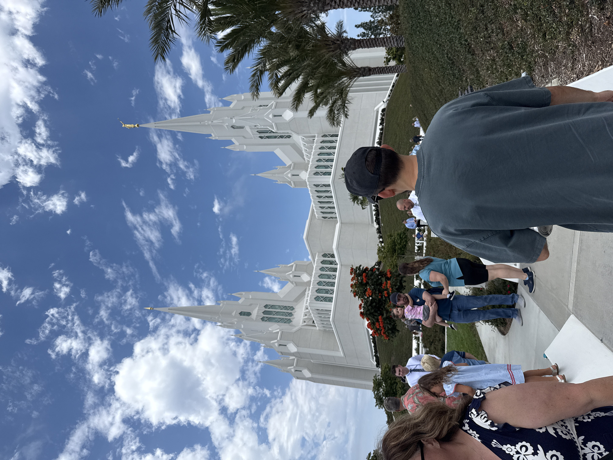 The image shows the San Diego California Temple of The Church of Jesus Christ of Latter-day Saints, located in San Diego, California, United States of America. It is a large, white, ornate temple with multiple spires, one of which is topped with a golden statue of the Angel Moroni. The temple is situated on a hill covered in green grass and landscaping. In the foreground, a group of people are walking on a paved pathway. Some are looking towards the temple, while others are looking at their phones or talking to each other. The sky is blue with white, fluffy clouds. The overall atmosphere is serene and majestic.