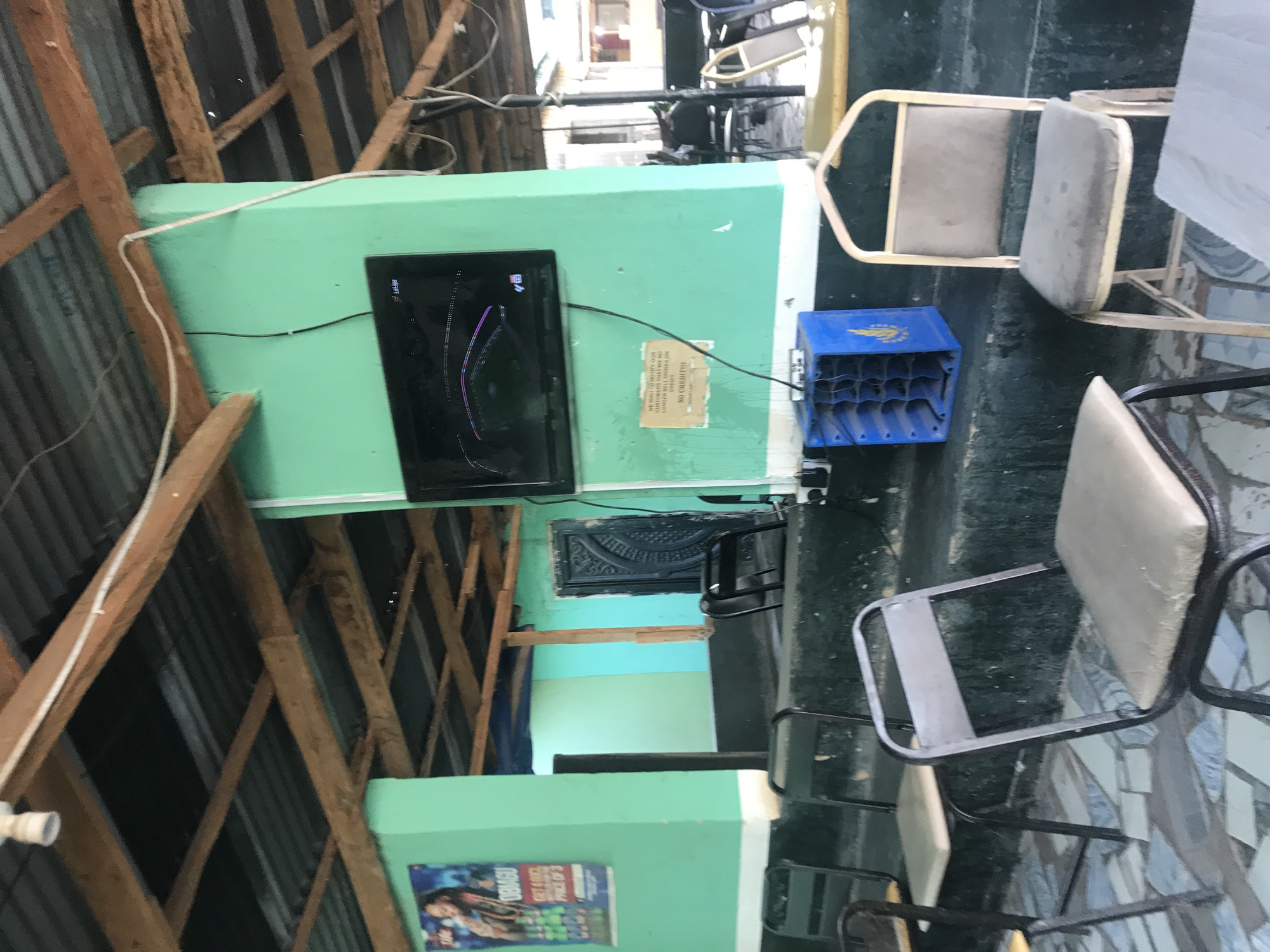 The image depicts an interior space with a corrugated metal roof supported by wooden beams, likely in New Stadium, Nigeria. A mint-green wall is visible, displaying a flat-screen TV and a sign. The sign reads "We wish to notify our customers that we no longer sell drinks on credit. No credit." Below the wall, there's a blue plastic crate. Several metal chairs with cushioned seats are scattered around a patterned tiled floor. A movie poster is also visible on another wall painted in a similar green hue.