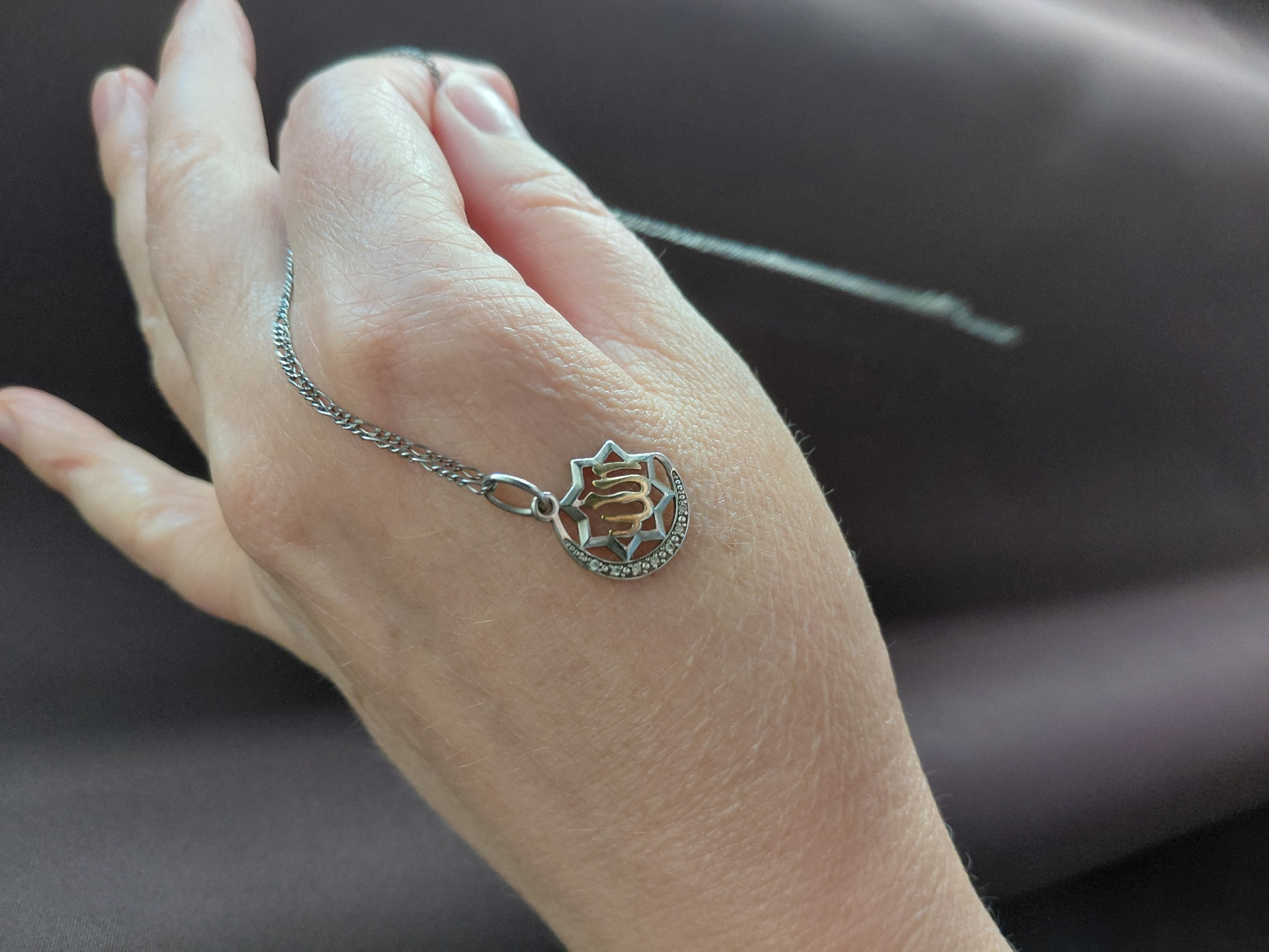 A close-up shot shows a hand holding a silver necklace with a crescent moon and star pendant. The pendant features Arabic calligraphy in gold, possibly a religious inscription, surrounded by small, clear stones. The hand and necklace are against a dark, textured fabric background, which could be a garment or a curtain. The lighting is soft and natural, casting gentle shadows. There is no discernible urban landscape or any elements that would indicate the location as Novosibirsk, Russia.