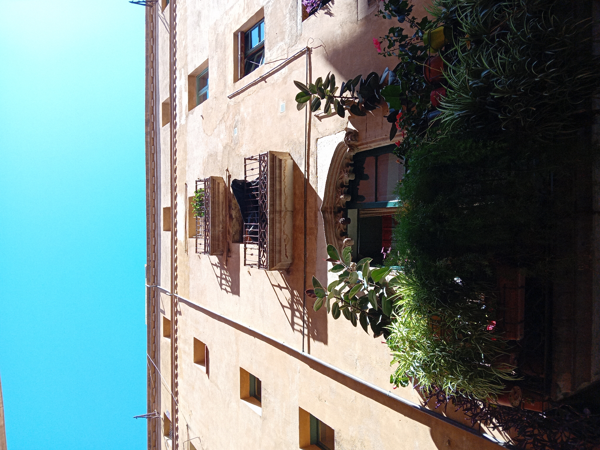 The image depicts a building facade in Barcelona, Spain. The building is multi-storied with light-colored, textured walls and several windows. Some windows have decorative iron grates, while others are framed by stone arches. Lush plants and flowers cascade from some of the windows, adding greenery to the urban scene. The sky above is a clear, bright blue. A vertical architectural detail runs along the edge of the building, accentuating its height. The image has a slightly tilted perspective, looking upwards at the building.
