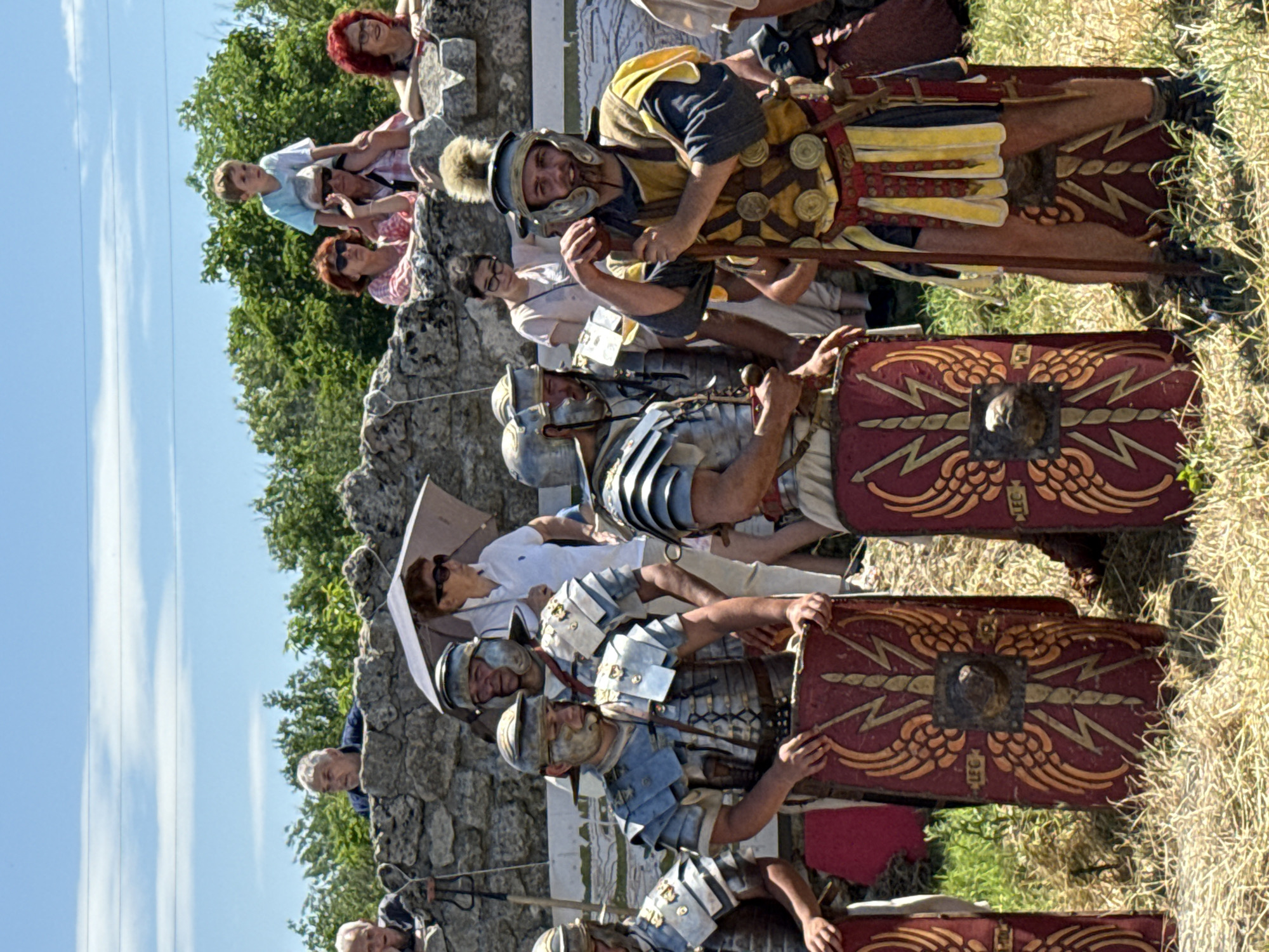 This image captures an outdoor historical reenactment in Gigen, Bulgaria, under a bright blue sky with faint utility lines visible at the top. The foreground features approximately six male individuals dressed as Roman soldiers. They wear various forms of metallic Roman armor, including segmented plates and chainmail, paired with helmets. One soldier on the far right, wearing a yellow and red tunic adorned with circular medallions and a distinctive plumed helmet, stands with a staff in his right hand, facing forward with a slight smile. The soldiers hold or rest large, rectangular scuta (shields) against their bodies; these shields are predominantly red with gold/yellow winged lightning bolt designs and a central metallic boss, some bearing the inscription "LEG". The ground beneath them is covered in dry, sparse grass.
Behind the Roman reenactors, a low, rough-hewn stone wall spans the midground, separating them from a group of spectators. Several people are visible observing from behind or on top of this wall. On the left side of the wall, an older man with grey hair, wearing a blue shirt, looks over. On the right side of the wall, a young boy with light hair sits on the shoulders of an older man wearing sunglasses. Adjacent to them, a woman with auburn hair and sunglasses holds a beige umbrella for shade. Further right along the wall, another woman with reddish hair leans forward, smiling. The spectators are dressed in modern, casual clothing. The background consists of dense green trees and foliage under the clear sky.