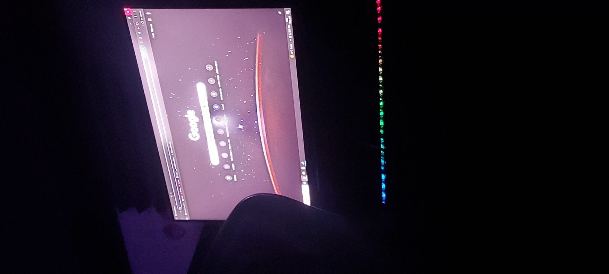 The image shows a computer monitor in a dark room. The screen displays a desktop with a Google search bar and other icons. To the right of the monitor, a vertical strip of colored lights, progressing through red, green, and blue, is visible. The lower part of a chair is also visible in the foreground, obscuring the bottom of the screen.