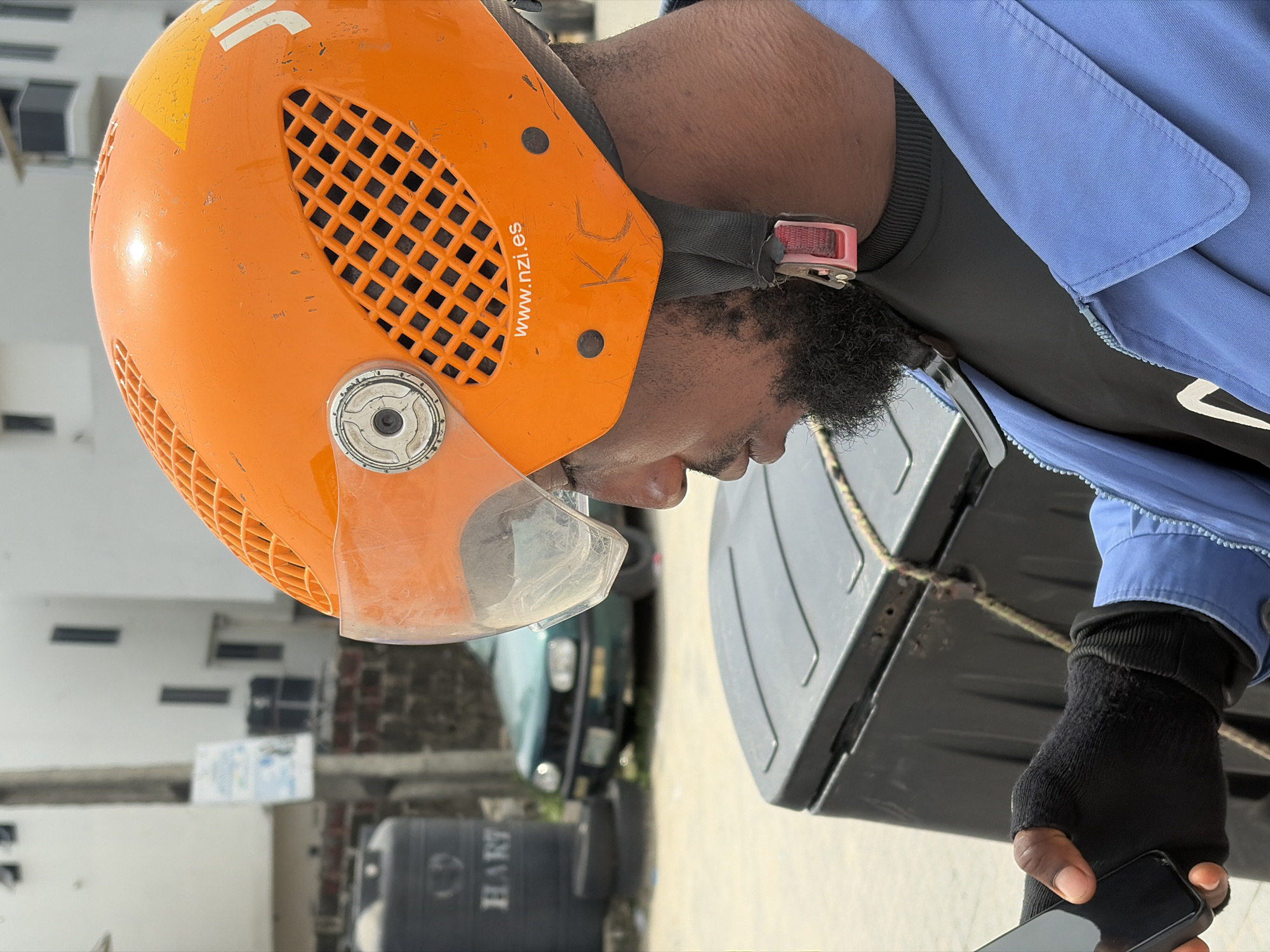 The image depicts a man wearing an orange helmet with visible ventilation holes and a clear visor. The helmet has markings including "www.nzi.es" and the letters "KC". He is also wearing a blue jacket over a black shirt and black fingerless gloves. The man appears to be holding a smartphone in his right hand. A gray container is partially visible in the foreground, secured with rope. The background includes a green car, a black water tank labeled "HART", and a building with white walls and dark windows. The scene is located in Igboefon, Nigeria.