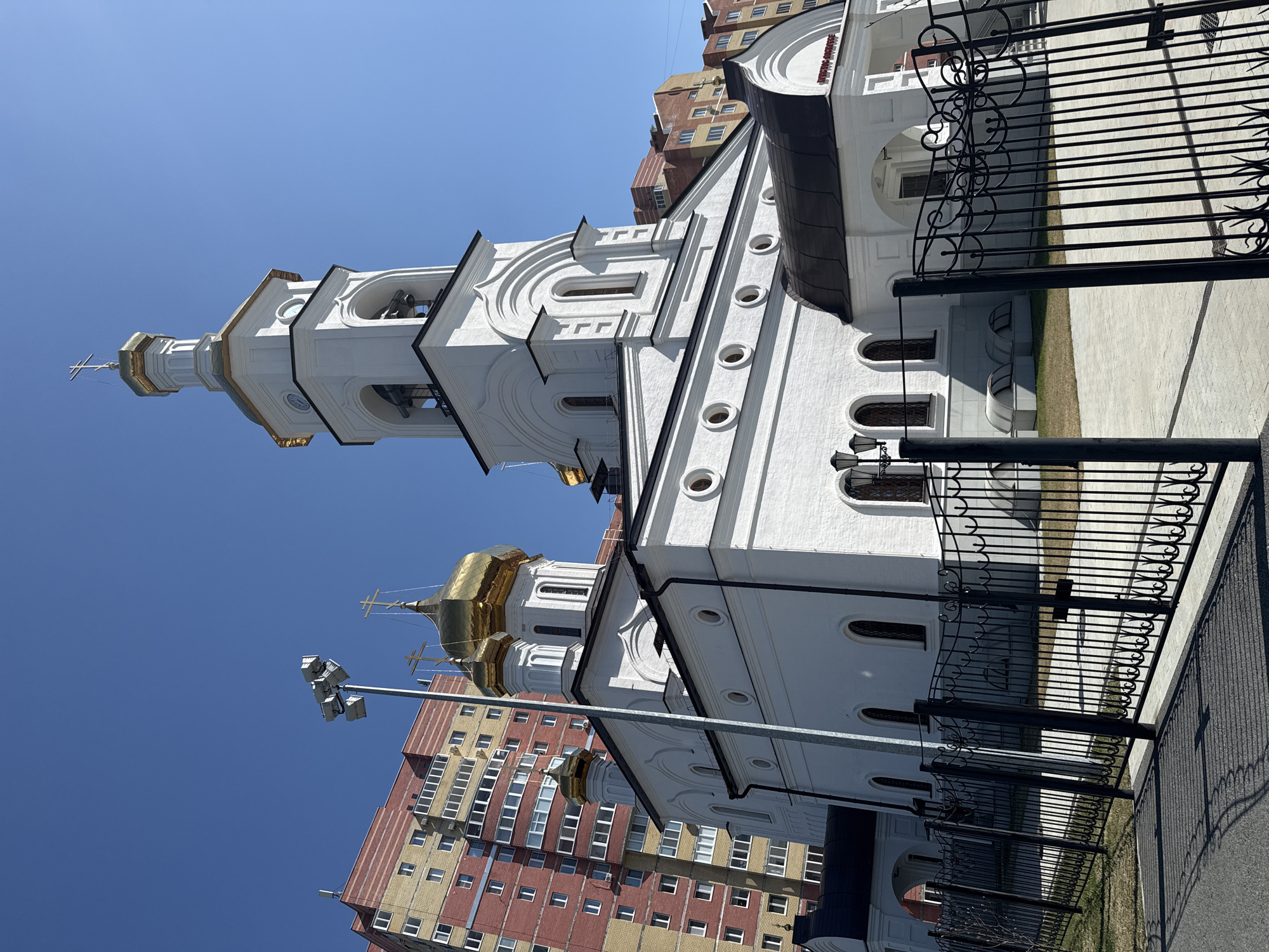 The image shows a white Orthodox church with golden domes, topped with crosses, located in Tobolsk, Russia. The church has a bell tower with arches and a tall, slender spire. A modern high-rise apartment building stands in the background, featuring a mix of red brick and yellow siding. A decorative black metal fence runs along the side of the church grounds. The sky is a clear, bright blue. A streetlight with multiple spotlights is positioned near the church. The image is taken from a low angle, looking upwards. The photograph appears to have been rotated.
