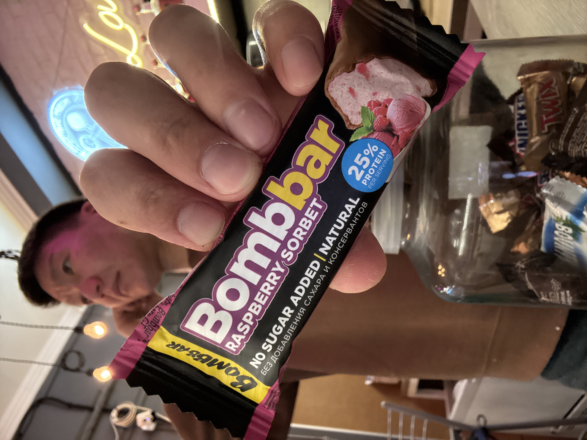 A person is holding a "Bombbar Raspberry Sorbet" protein bar. The packaging indicates "No Sugar Added | Natural" and "25% Protein Per Serving" along with some Russian text. A jar of candy bars, including Snickers and Twix, is visible in the background. The person appears to be indoors with neon signs and other lighting elements visible. "Bombbar" is also visible on a second yellow and pink packaging. I can not determine if the image was taken in Khimki, Russia.