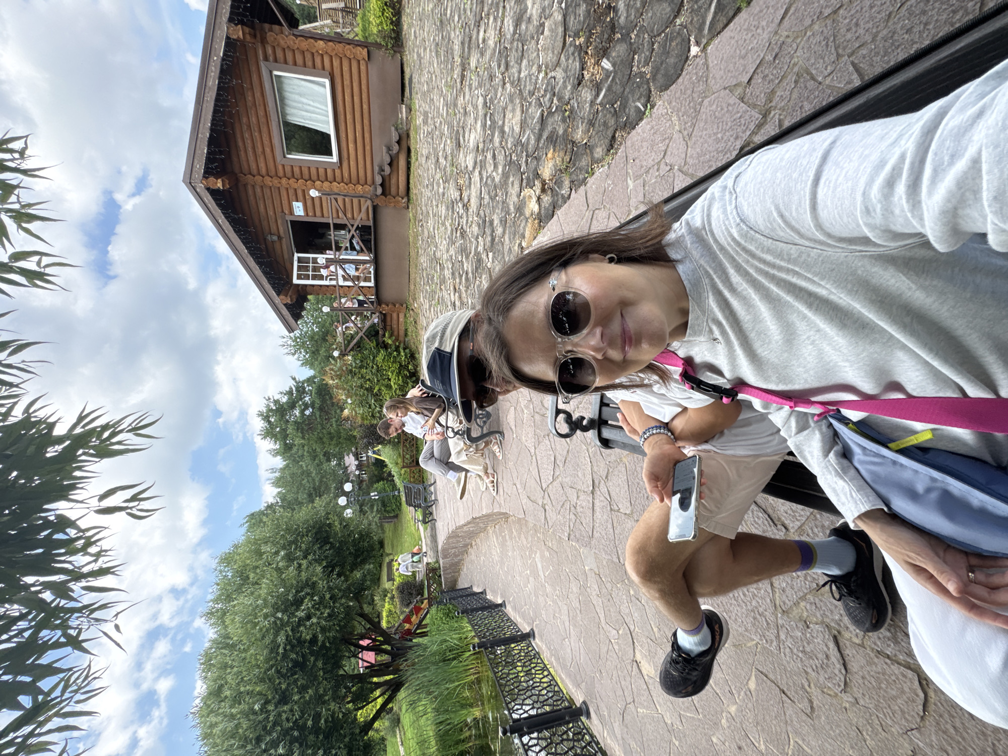 A selfie is taken from a seated perspective on a bench, focusing on a woman in the foreground. She wears sunglasses and a grey sweatshirt, with a pink strap and blue bag visible. Her left hand rests on her lap, displaying a ring. To her right, a man is seated, wearing a hat, sunglasses, and a light-colored shirt. He holds a smartphone in his right hand. Both are on a bench with a dark frame.
In the background, a cobblestone path curves to the left, bordering a body of water with a decorative fence. Further back, a rustic wooden cabin with a porch and a sign is visible. Several people are seated on benches in the middle ground, and others are walking around the area. Trees and greenery surround the scene, with a cloudy sky overhead. The overall setting appears to be an outdoor recreational area. This location is likely in Izhevsk, Russia, given the context.