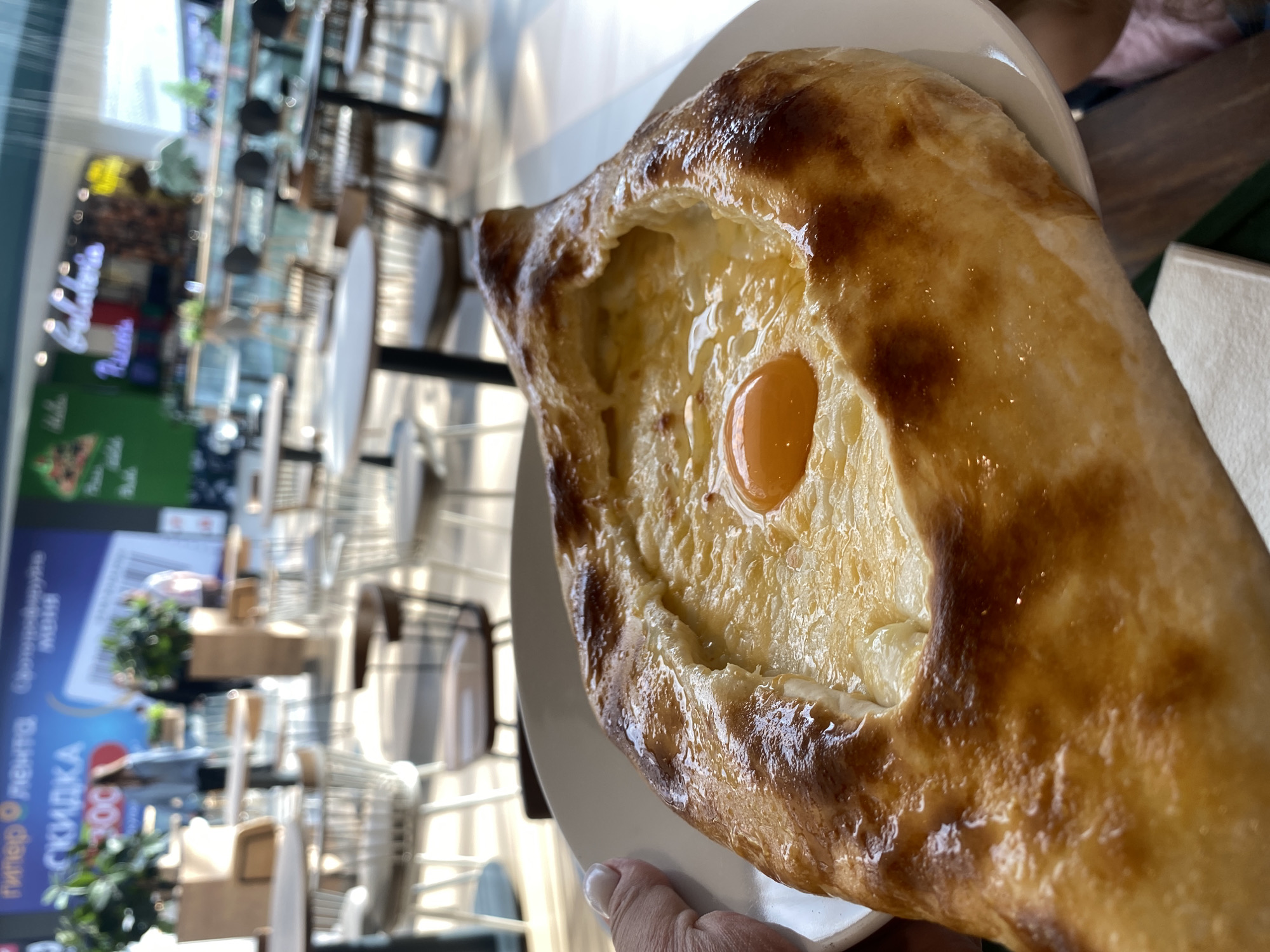 The image shows a close-up of an Adjaruli Khachapuri, a traditional Georgian bread dish, served on a white plate. The bread is boat-shaped with a golden-brown crust and filled with melted cheese and a raw egg yolk in the center.
In the background, out of focus, is a seating area with tables and chairs, suggesting a restaurant or cafe setting. A sign with Russian writing is visible, possibly indicating a promotion or advertisement. The location is likely in Perm, Russia, as indicated in the prompt. The environment appears to be indoors, with natural light coming through the windows, creating a bright and airy atmosphere. Someone is holding the plate.
