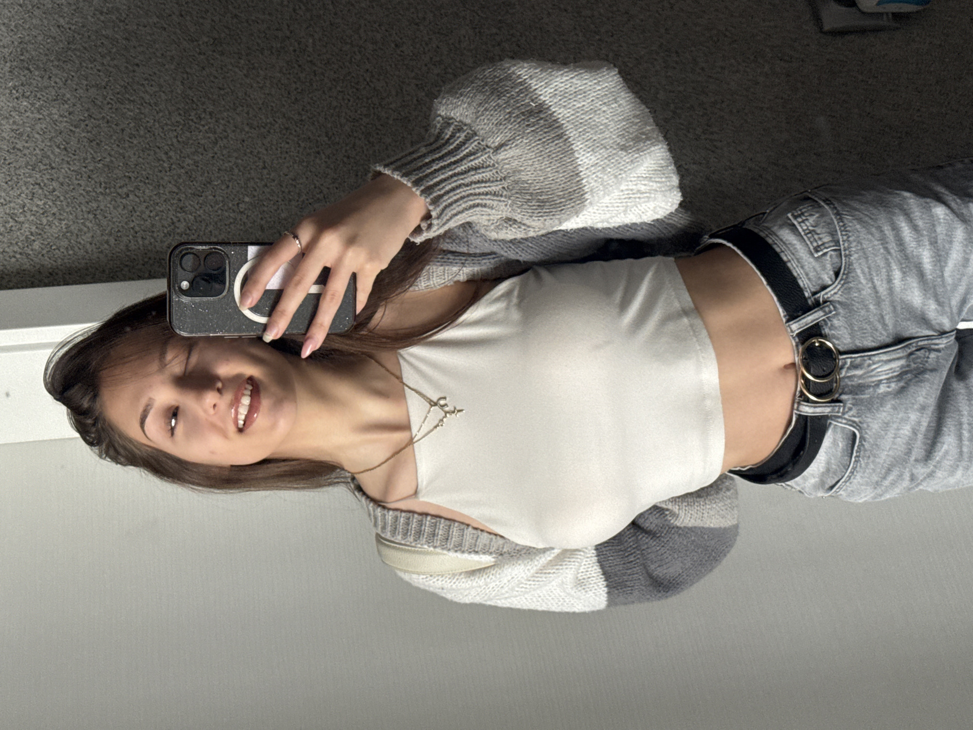 A young woman is taking a selfie. She has light brown hair, a white tank top, a gray and white cardigan, and gray jeans with a black belt. She is wearing a ring on her left hand and a necklace. She is holding a black smartphone. The background is a gray carpet and a light gray wall. The location cannot be determined from the image.