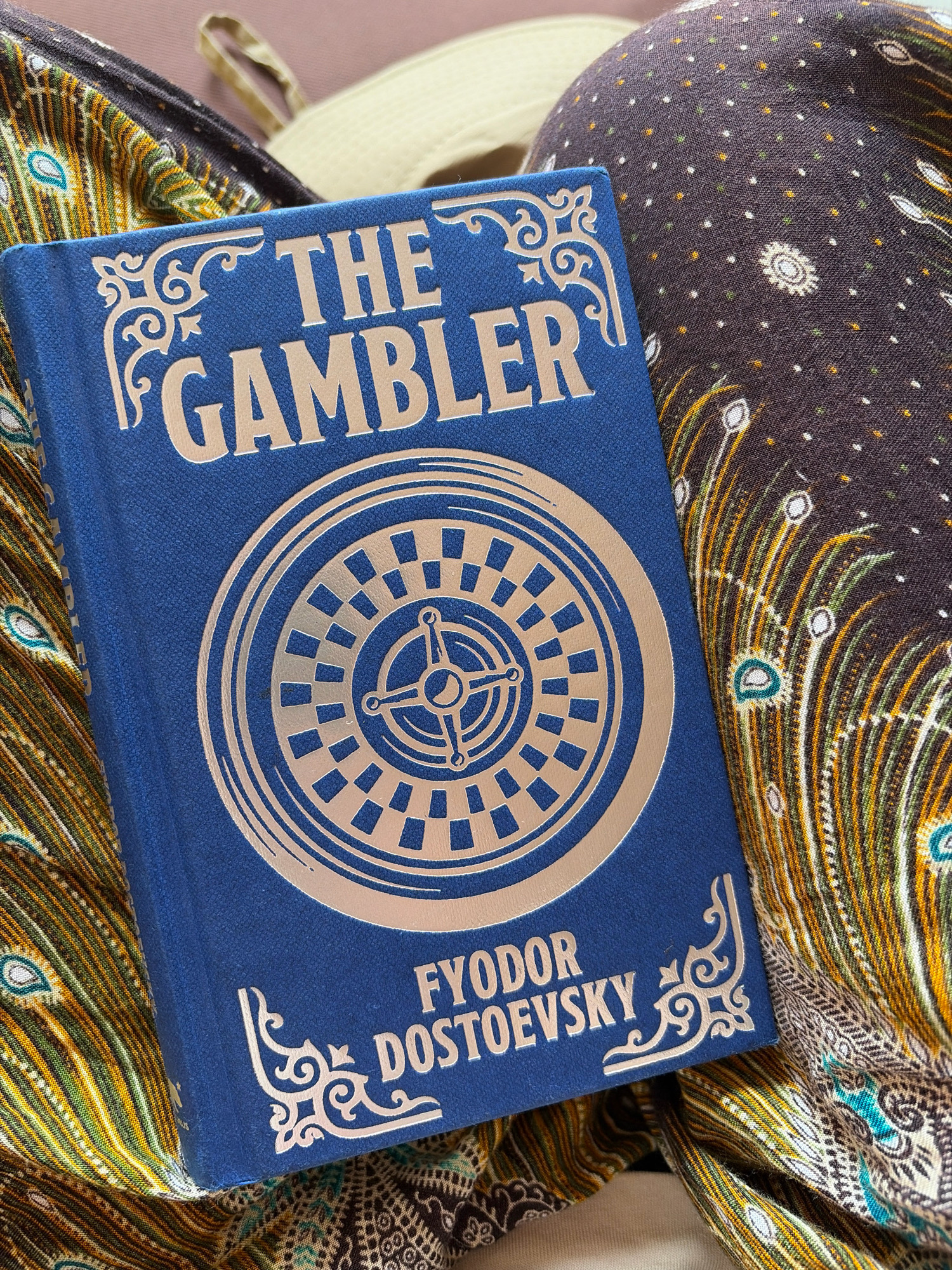 A blue hardcover book titled "THE GAMBLER" by FYODOR DOSTOEVSKY lies open on a patterned fabric. The book's cover features a roulette wheel design in a metallic silver finish, with decorative scrollwork at the top and bottom corners. The fabric underneath displays a vibrant pattern of peacock feather motifs in shades of green, yellow, brown, and blue. The book is angled, resting on its spine and partially open, revealing some of the pages. The background is blurred, suggesting a shallow depth of field. The scene is indoor, with soft lighting. There is no discernible location in Lalitpur, Nepal.