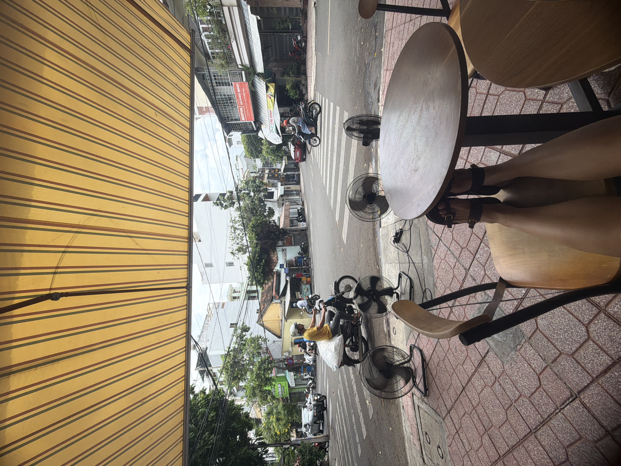The scene captures a street view from a cafe in Nha Trang Ward, Vietnam. The yellow-striped awning of the cafe dominates the left side of the frame. In the center, a street is visible, populated with motorbikes and pedestrians moving along the crosswalk and road. Several buildings and trees line the street, typical of an urban environment. On the right side of the frame, there are two circular tables, one occupied by a person whose legs and feet in sandals are visible. Three electric fans are seen nearby on the pavement. The image appears to be taken during the daytime.