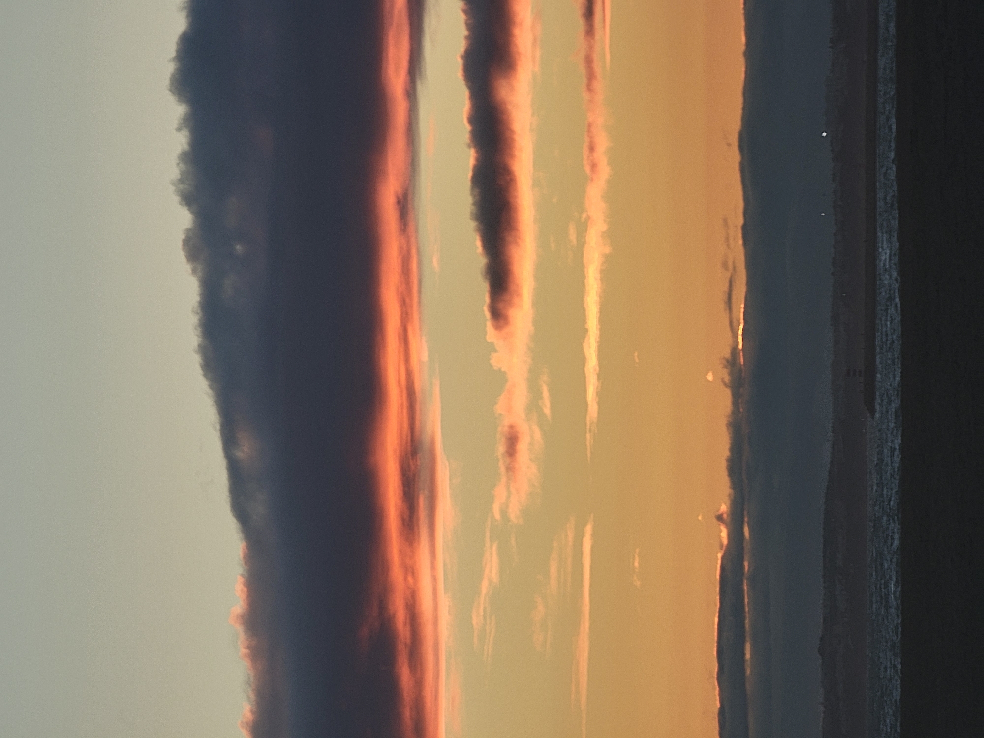 The image depicts a coastal scene at dusk, likely Quarteira, Portugal, characterized by a layered sky of muted oranges, yellows, and blues, intersected by bands of dark, wispy clouds. The upper portion of the sky is a pale, almost greyish-blue, transitioning to warmer tones as it descends. A prominent, dark, horizontal cloud formation stretches across the mid-upper section, tinged with streaks of coral and pink along its lower edge. Below this, lighter, scattered clouds reflect the fading sunlight in soft hues of peach and gold. The horizon is marked by a dark, silhouetted landmass, suggesting a coastline with distant, indistinct structures and possibly a lighthouse on the right. A body of water, appearing choppy with whitecaps, occupies the lower third of the frame, reflecting the twilight colors. The foreground is a dark, featureless expanse of what appears to be a beach or sand, devoid of any discernible objects or activity. The overall atmosphere is tranquil and serene, indicative of the end of a day.