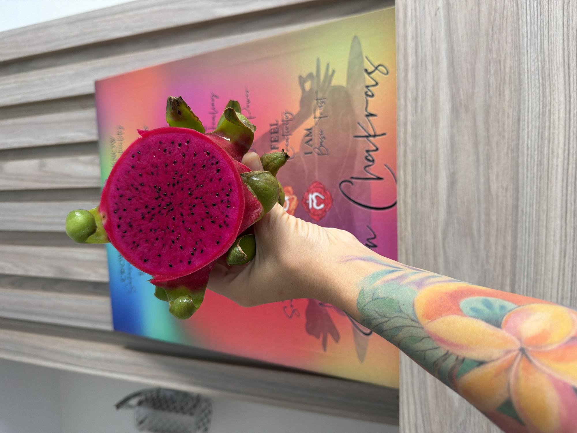 A person's left forearm, adorned with a vibrant floral tattoo in shades of yellow, orange, pink, and green, is held up, supporting a halved dragon fruit. The fruit's interior is a striking magenta with numerous small black seeds. Behind the fruit and arm is a book or poster with a gradient of colors, transitioning from blue and green to pink and yellow, featuring the word "Chakras" in cursive script and other text. The background consists of vertical wooden slats. This image was captured on a wooden surface, possibly a table or shelf. The lighting suggests it is daytime. The image does not contain any geographical indicators of Nha Trang, Vietnam.