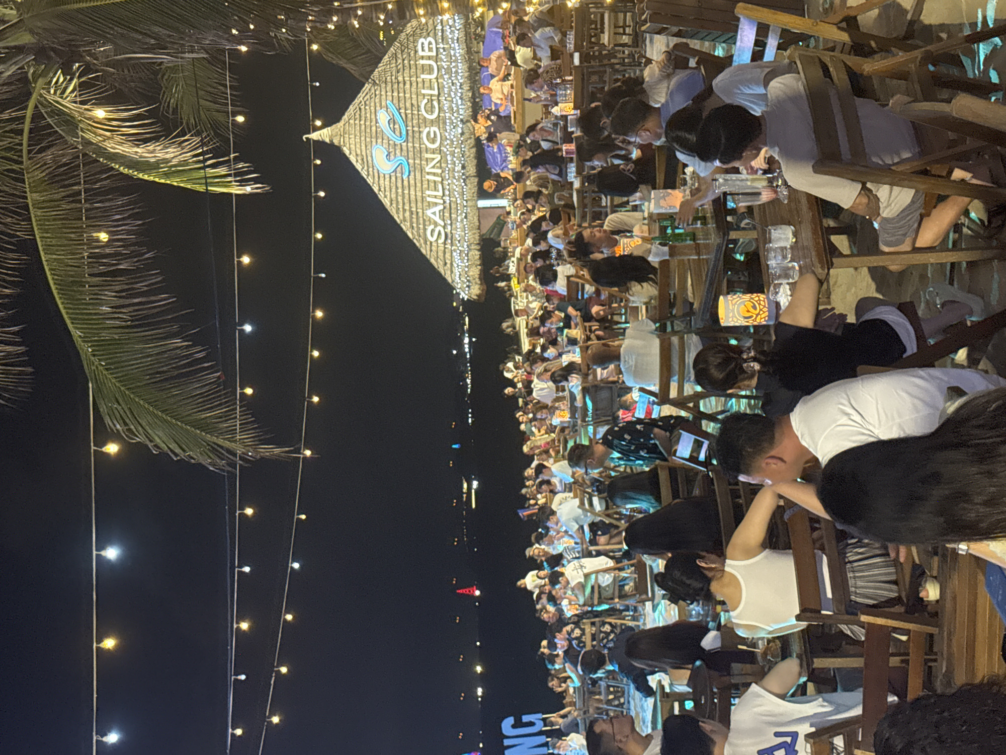 Here is a detailed description of the image:
**Location:** The image shows an outdoor venue filled with people, presumably in a city of Unknown, Unknown. A prominent structure is labeled "Sailing Club." The area is illuminated by string lights, which hang in rows throughout the scene.
**People:** The scene is densely populated with numerous individuals. They appear to be mostly seated at tables, possibly dining or socializing. Some are looking at their phones, while others seem to be engaged in conversations or eating.
**Subjects and Objects:** The primary subjects are the people occupying the venue. Notable objects include the tables and chairs at which they are seated, glassware (possibly containing drinks), and personal electronic devices (phones).
**Actions and Interactions:** The primary activity is people engaging in social interactions, dining, and using their personal electronic devices.
**Environment:** The environment includes the outdoor venue itself, the “Sailing Club” structure, and the surrounding ambient light in the evening or nighttime setting. There is a palm tree in the frame as well.