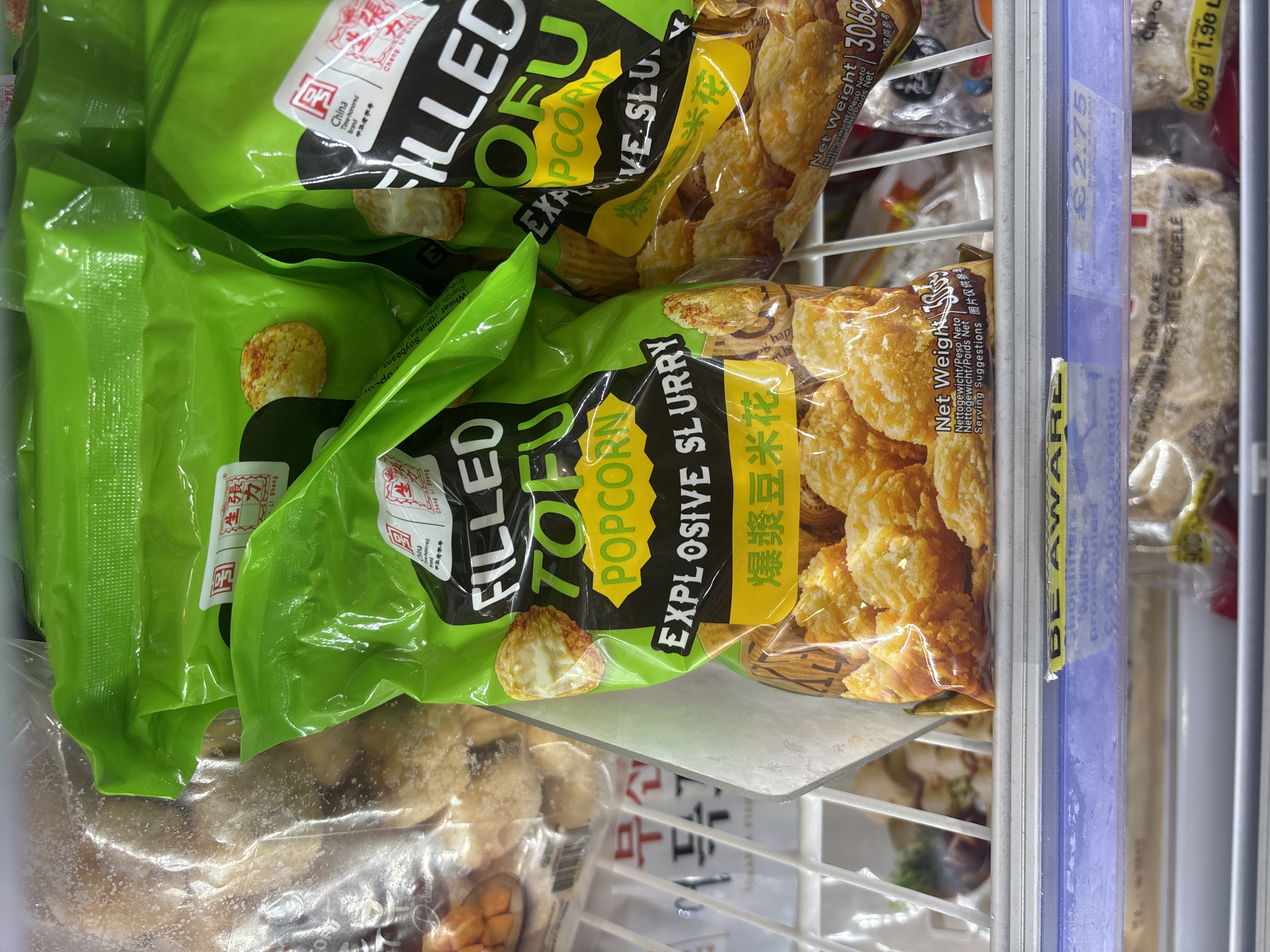 The image shows bags of frozen "FILLED TOFU" popcorn explosive slurry in a freezer, likely in a grocery store in London, United Kingdom. The packaging is green and yellow, with Chinese characters and English descriptions. The price tag visible is £2.75 for a different product ("DE POISISON PRE-RITE CONGELE"). The items are arranged on shelves within the freezer, with other frozen products visible nearby.
