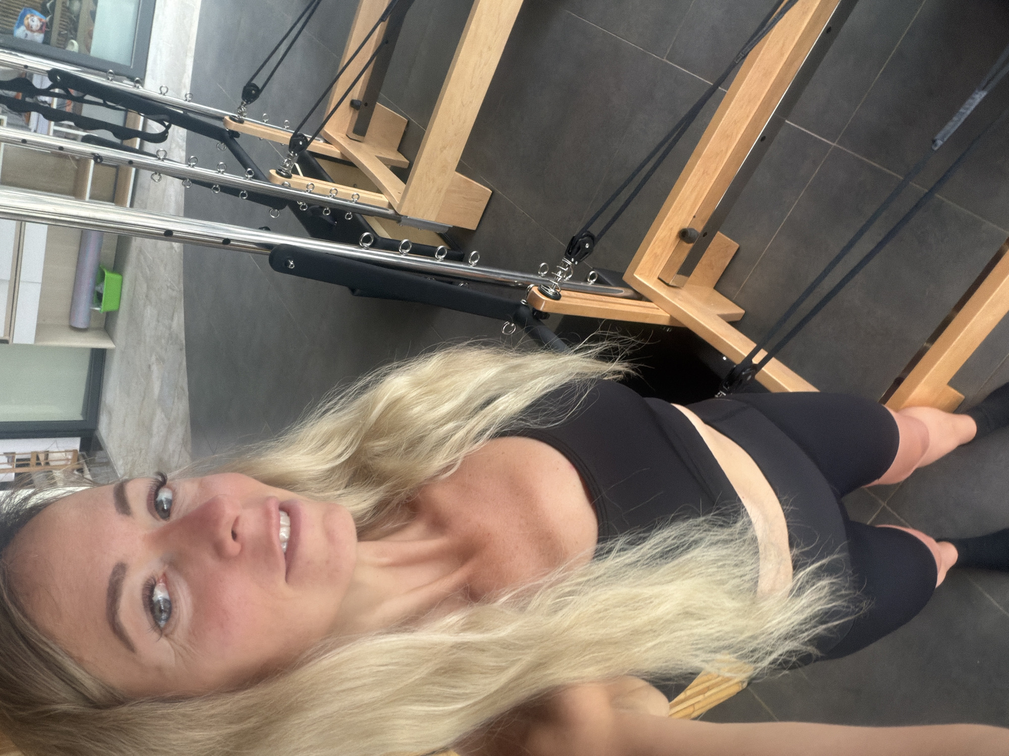 A blonde woman stands in a Pilates studio, looking into the camera. She is wearing a black sports bra and black bike shorts, with black socks on her feet. The studio has dark grey tiled floors and a large wooden and metal Pilates reformer machine. The reformer has a wooden frame, metal bars, and black straps and pulleys. In the background, there are shelves with exercise equipment, including a green bucket and a rolled-up mat. The woman's left arm is slightly extended towards the camera.